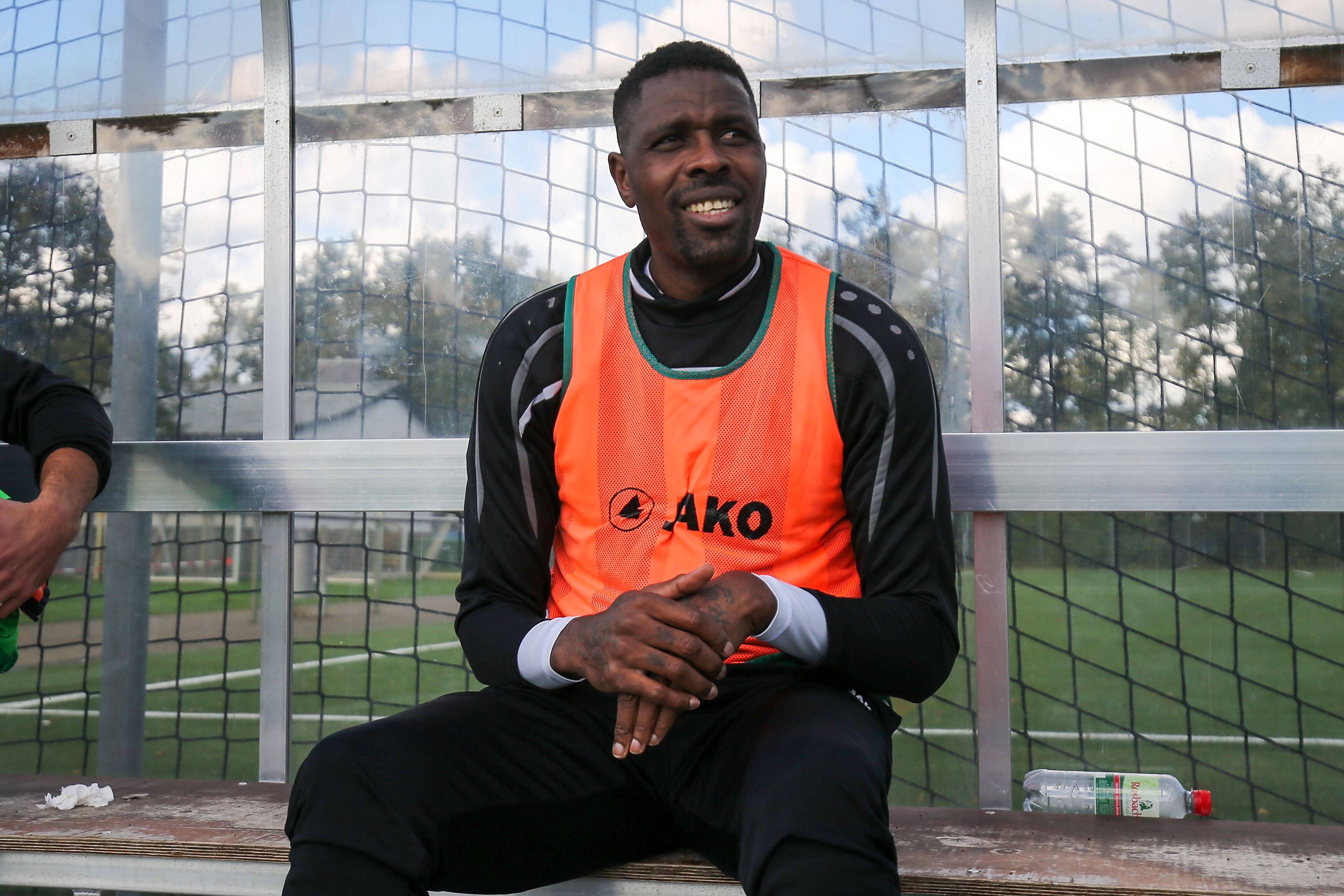 Ex-Gladbacher Mo Idrissou sitz am 10. Oktober 2020 im orangenen Leibchen beim Spiel gegen den Hunfelder SV auf der Ersatzbank beim SC Viktoria Griesheim und lächelt.