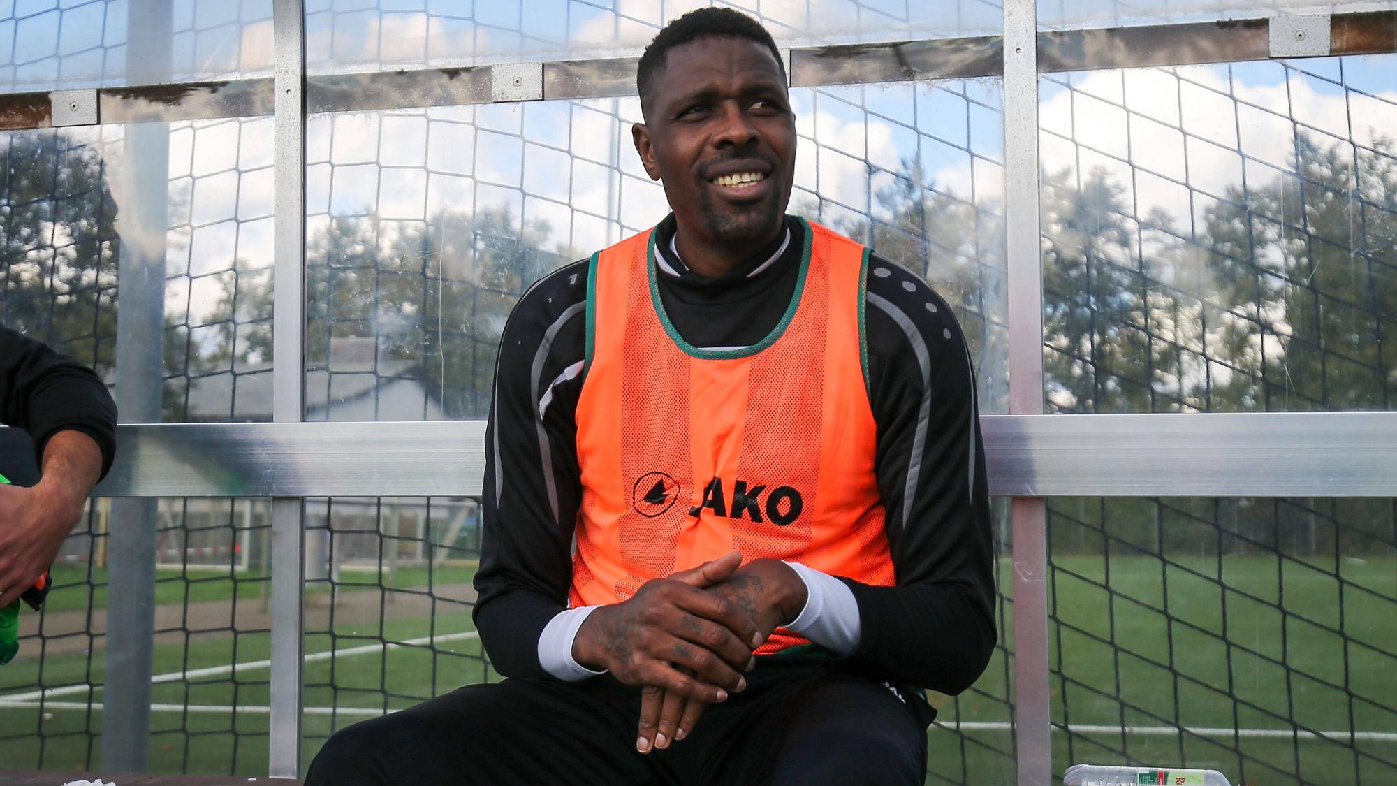 Ex-Gladbacher Mo Idrissou sitz am 10. Oktober 2020 im orangenen Leibchen beim Spiel gegen den Hunfelder SV auf der Ersatzbank beim SC Viktoria Griesheim und lächelt.