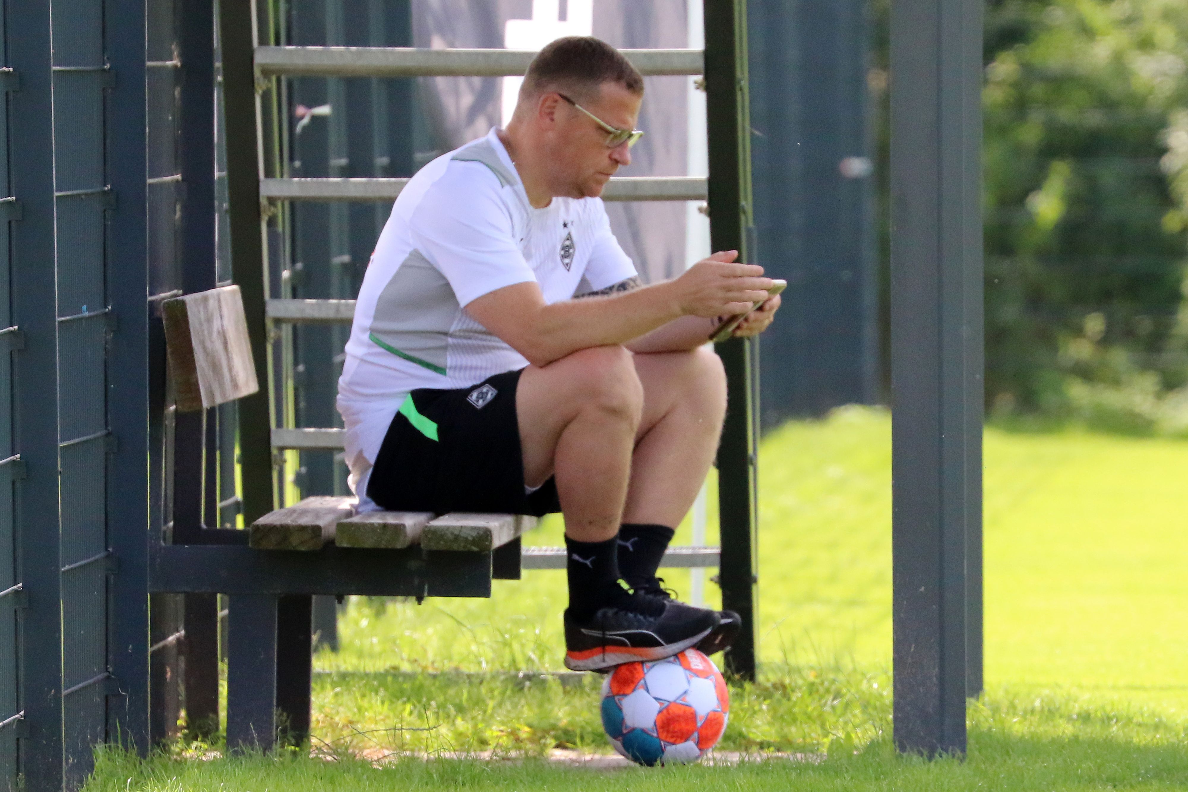 Gladbach-Manager Max Eberl sitzt auf einer Bank, derweil schaut er auf sein Smartphone. Seine Füße ruhen auf einem Fußball.