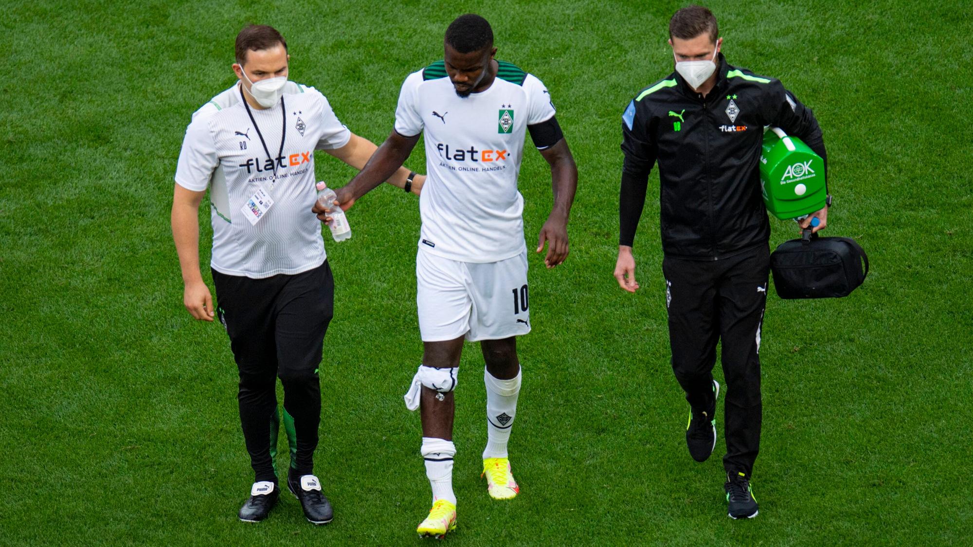 Gladbach-Stürmer Marcus Thuram (Mitte) humpelt beim verlorenen Rheinland-Duell (0:4) der Borussia in Leverkusen (21. August 2021) frühzeitig verletzt vom Platz. Er wird von zwei Betreuern, die eine Maske tragen, begleitet.