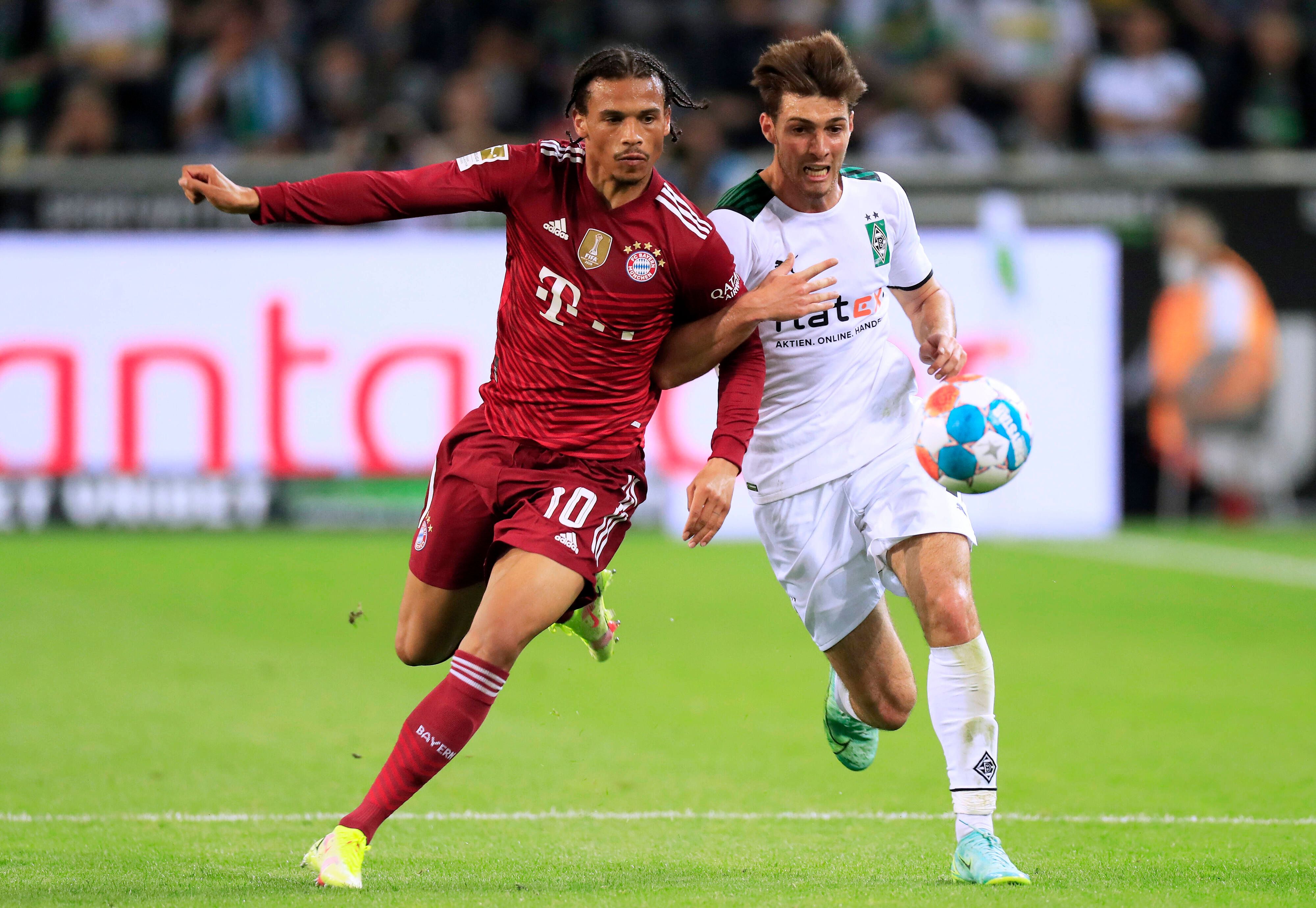 Verteidiger Joe Scally (r.) von Borussia Mönchengladbach im Laufduell mit Leroy Sane (l.) von Bayern München im Bundesliga-Duell am 13. August 2021. Beide Spieler haben den Ball im Blick, Scally hat sich mit seinem rechten Arm bei Sane eingehakt.