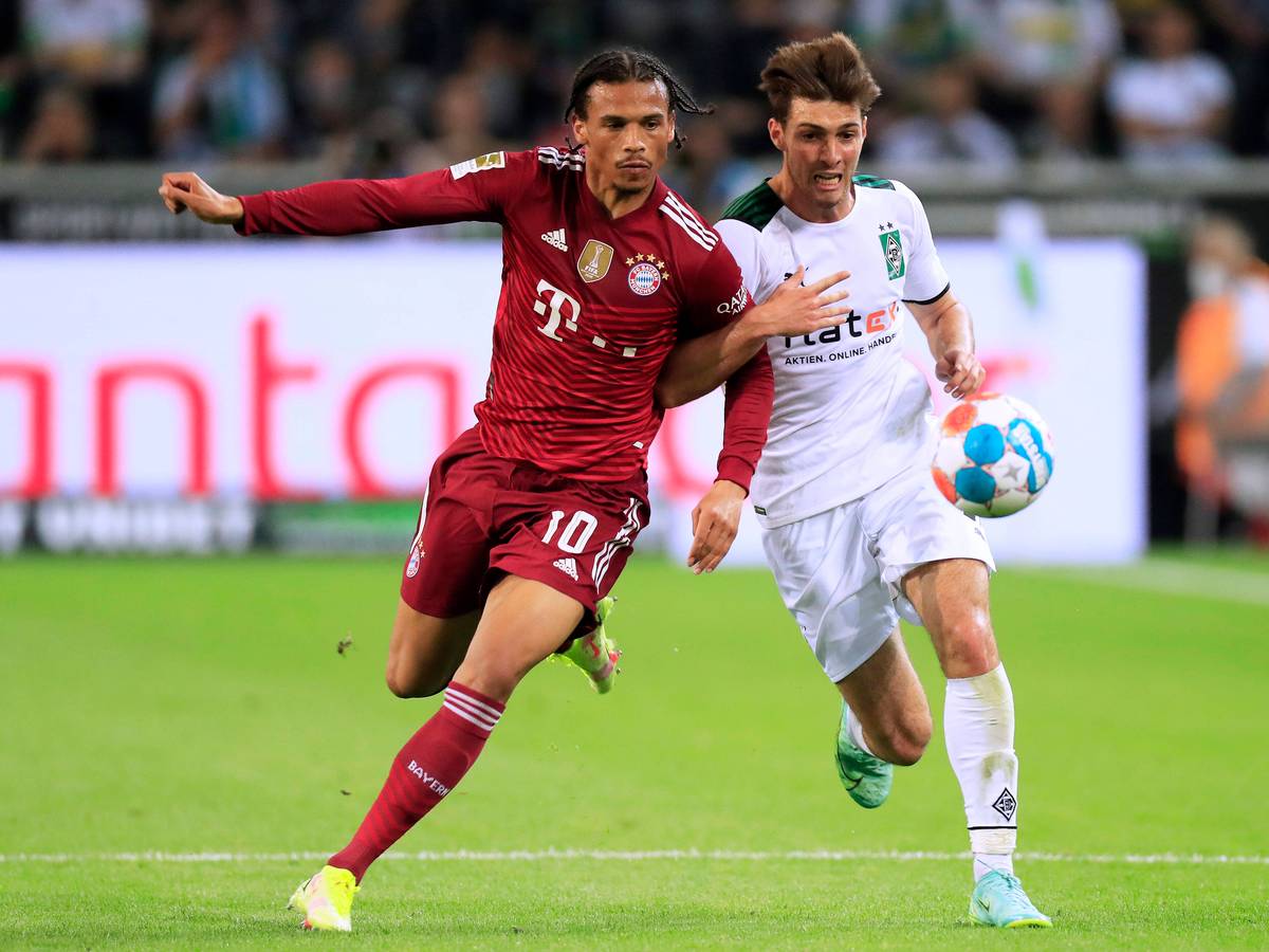 Verteidiger Joe Scally (r.) von Borussia Mönchengladbach im Laufduell mit Leroy Sane (l.) von Bayern München im Bundesliga-Duell am 13. August 2021. Beide Spieler haben den Ball im Blick, Scally hat sich mit seinem rechten Arm bei Sane eingehakt.