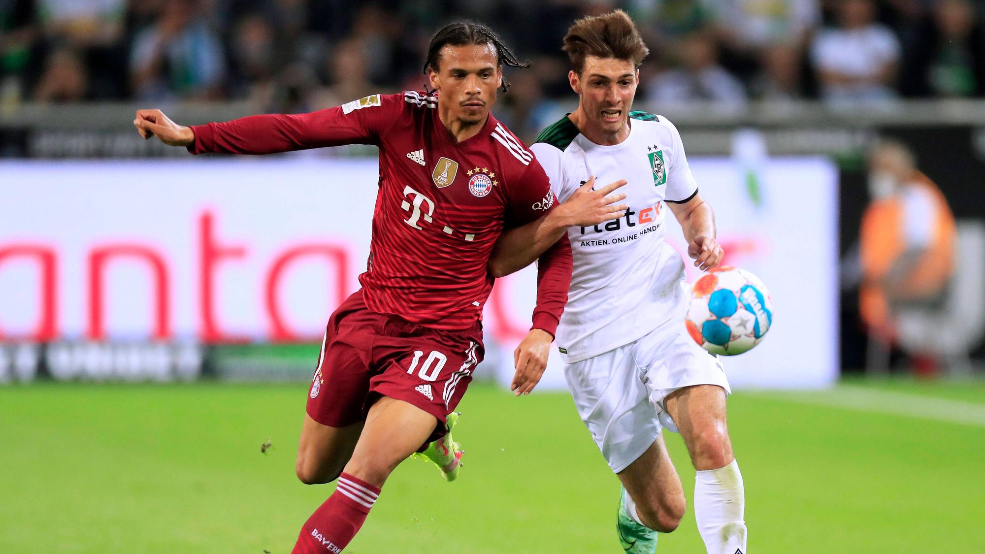 Verteidiger Joe Scally (r.) von Borussia Mönchengladbach im Laufduell mit Leroy Sane (l.) von Bayern München im Bundesliga-Duell am 13. August 2021. Beide Spieler haben den Ball im Blick, Scally hat sich mit seinem rechten Arm bei Sane eingehakt.