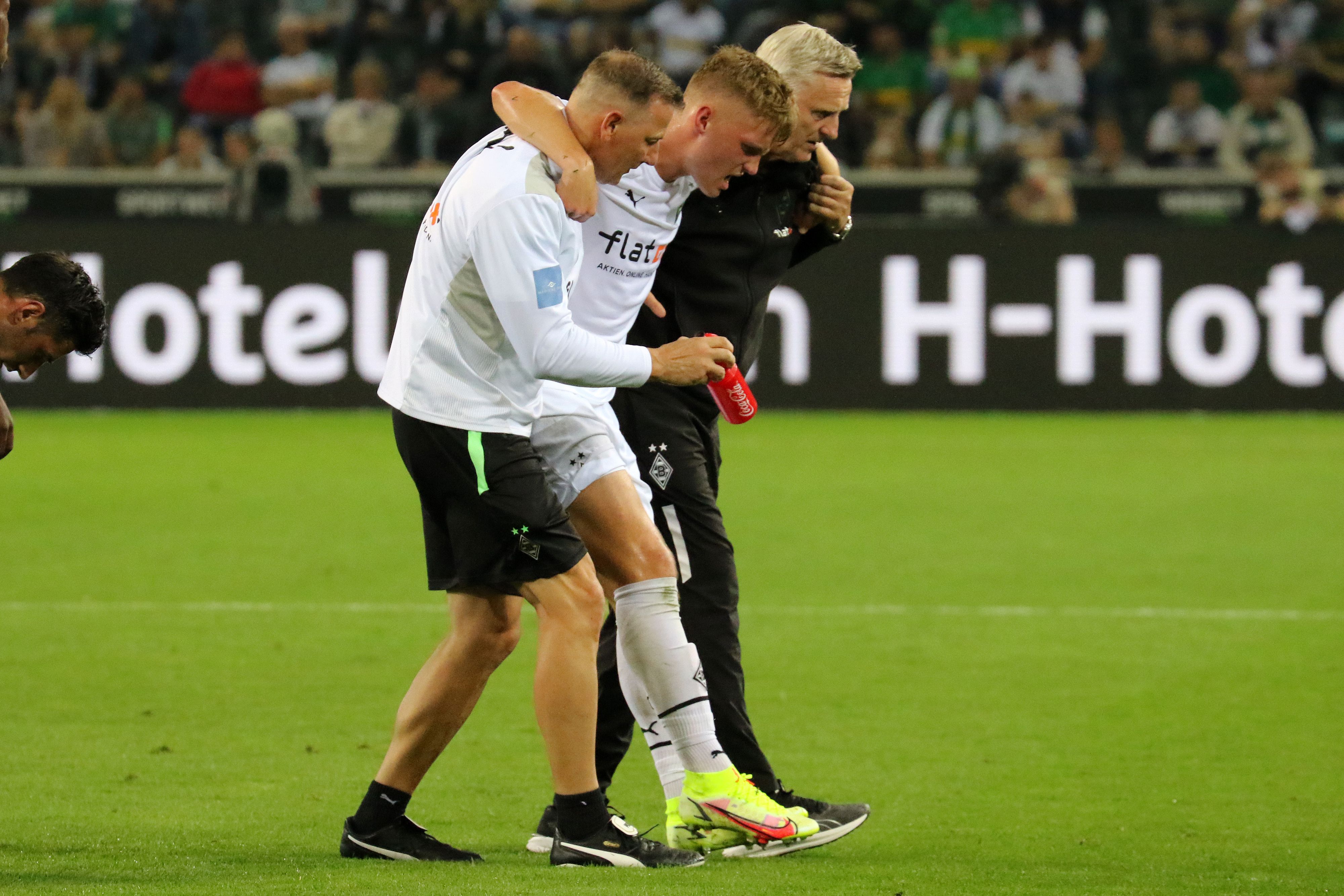 Luca Netz (M.), Spieler von Borussia Mönchengladbach, muss beim Bundesliga-Duell gegen Arminia Bielefeld (12. September 2021) verletzt vom Platz. Physio Dirk Müller (l.) und Team-Arzt Stefan Hertl (r.) stützen den Außenbahnspieler.