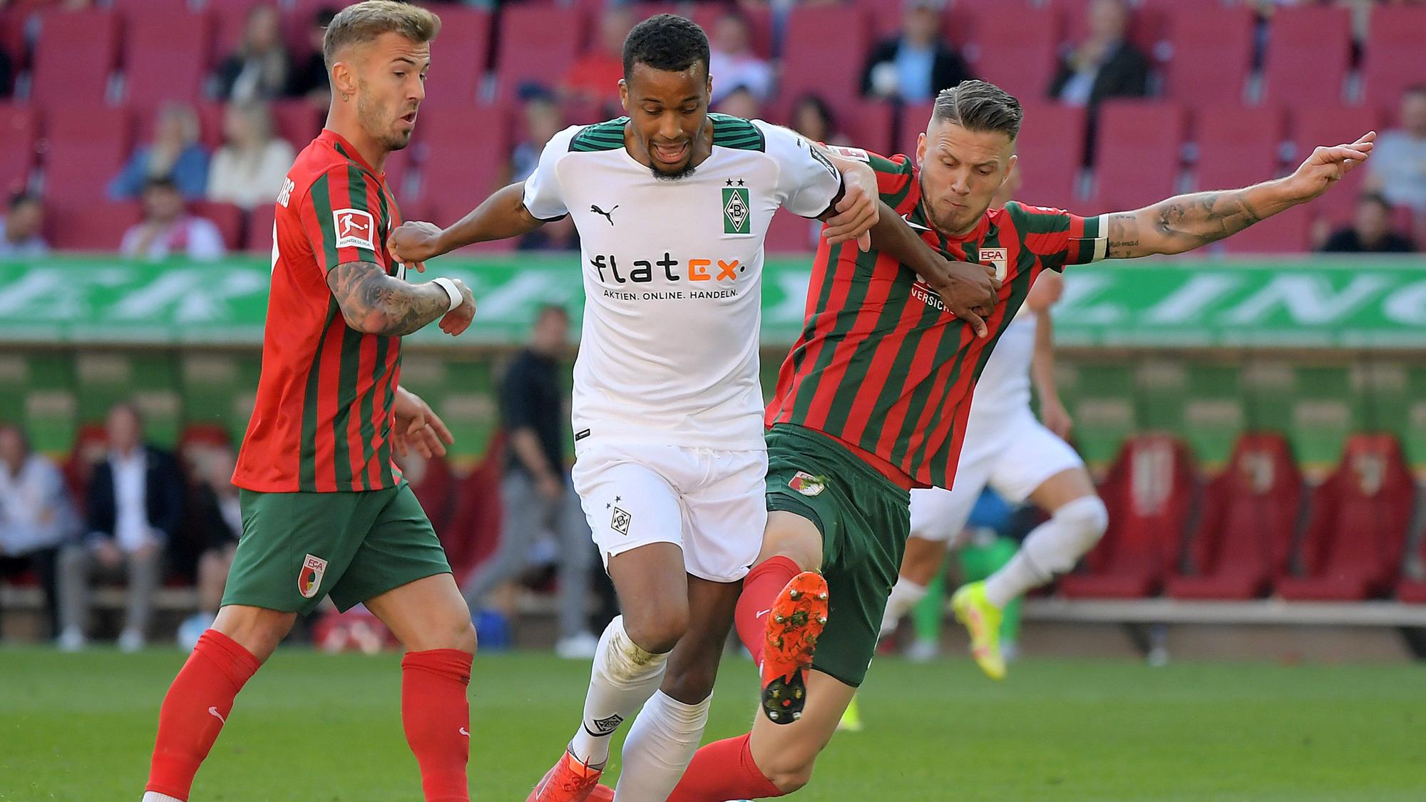 Alassane Plea von Borussia Mönchengladbach versucht seine beiden Augsburger Gegenspieler mit ausgestreckten Armen von sich wegzuhalten im Spiel gegen den FCA am 18. September 2021.