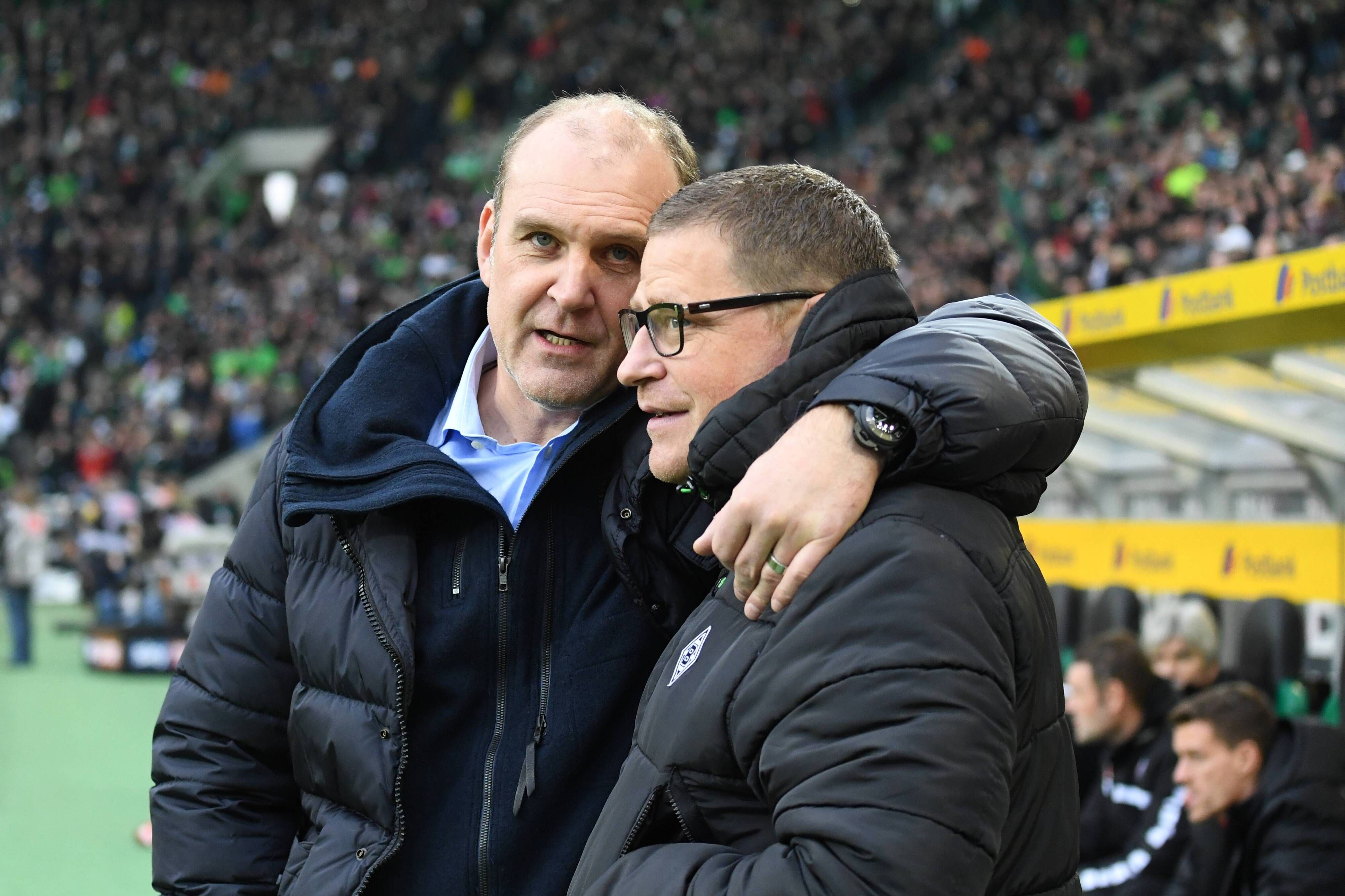 Jörg Schmadtke (l.), damals noch in Diensten des 1. FC Köln, umarmt am 19.11.2016 im Borussia-Park seinen Kumpel und Gladbach-Manager Max Eberl (r.) und schaut dabei in die Kamera eines Fotografen.