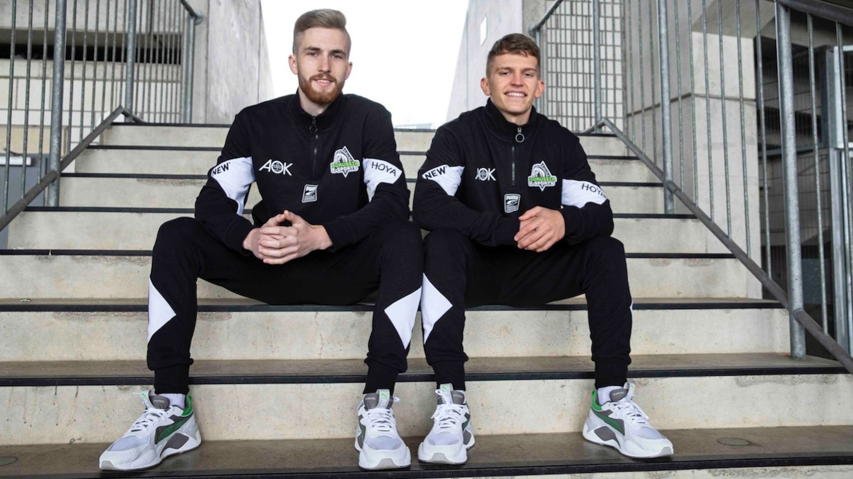 Borussias E-Sportler Yannick Reiners (l.) und Eleftherios „Lefti“ Ilias (r.), sitzen nebeneinander auf den Stufen im Borussia-Park und peilen die Meisterschaft an.