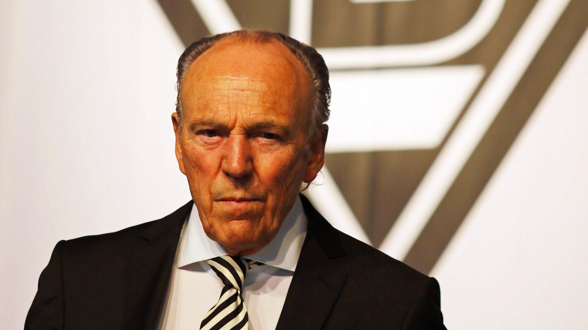 Gladbach-Präsident Rolf Königs steht vor einer Borussia-Fahne und blickt mit ernster Miene in die Kamera.