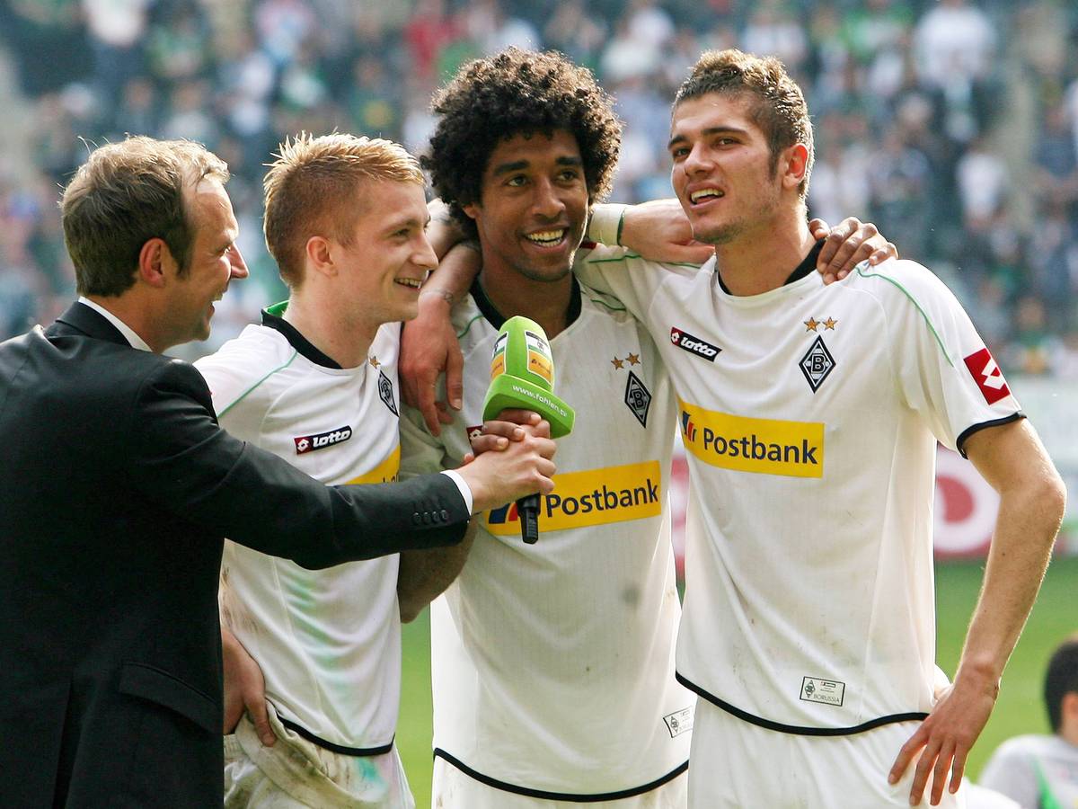 Stadionssprecher Torsten Knippertz im Gespräch mit Marco Reus, Dante und Roman Neustädter (v.l.n.r.) im Borussia-Park. Das Foto stammt vom 28. April 2012, die drei Spieler haben Borussia Mönchengladbach am Ende der Saison 2011/2012 verlassen. Reus, Dante und Neustädter umarmen sich, derweil hält Knippertz ihnen ein Mikrofon hin.
