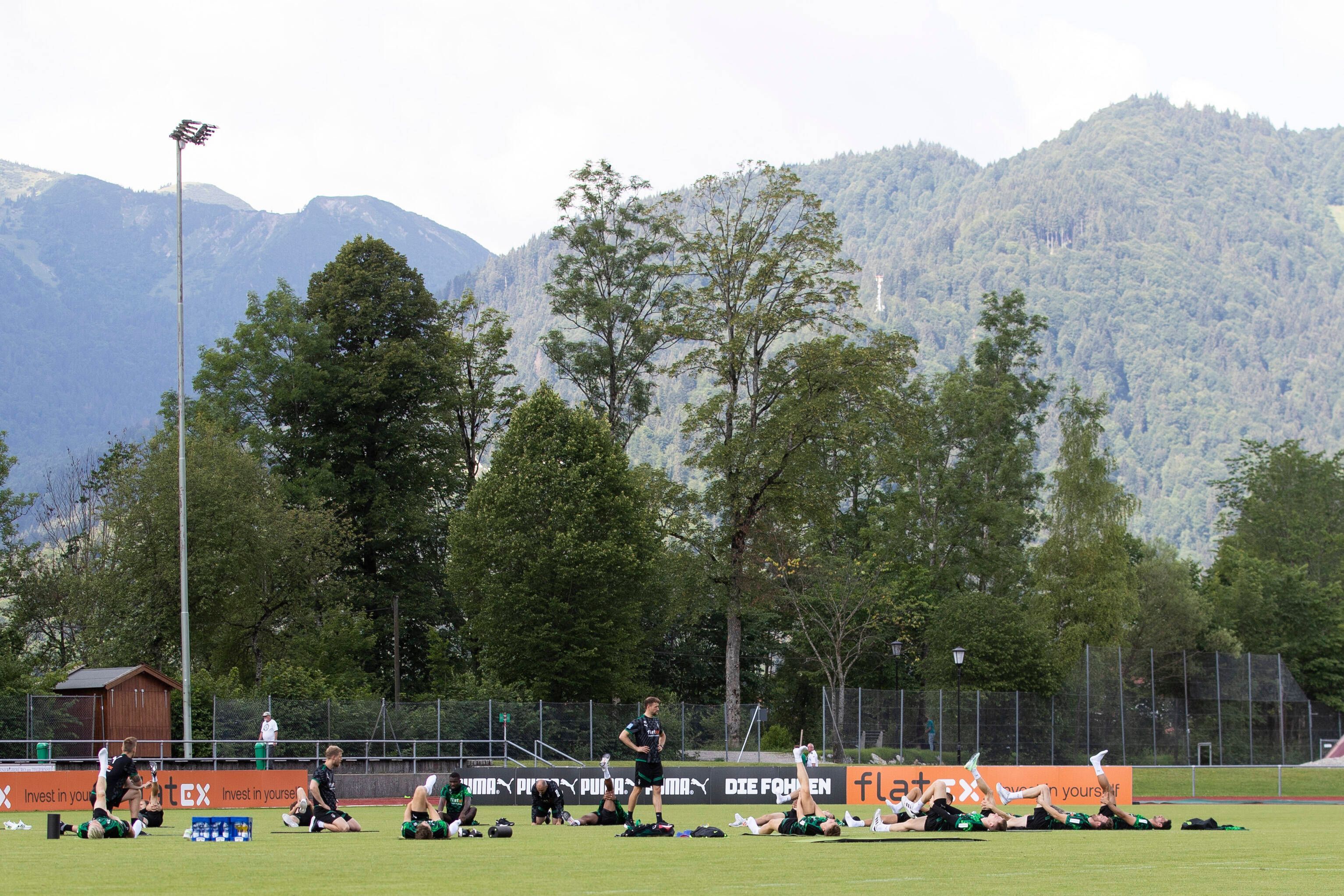 Die Gladbach-Profis im Trainingslager (6. Juli 2022) am Tegernsee in Rottach-Egern. Im Hintergrund sind die Bayerischen Alpen zu sehen. Die Spieler liegen auf dem Rasen und machen Stretching.