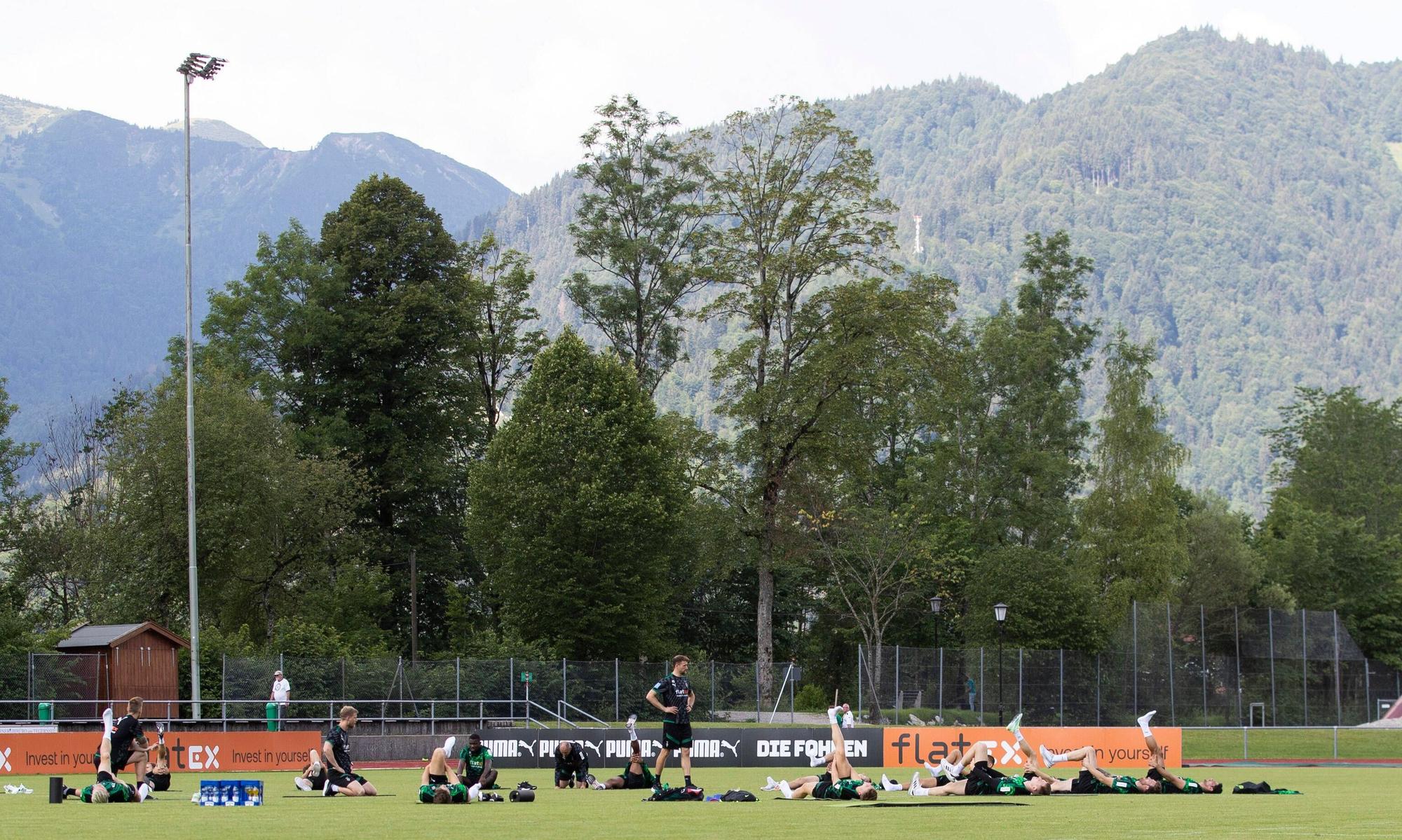 Die Gladbach-Profis im Trainingslager (6. Juli 2022) am Tegernsee in Rottach-Egern. Im Hintergrund sind die Bayerischen Alpen zu sehen. Die Spieler liegen auf dem Rasen und machen Stretching.