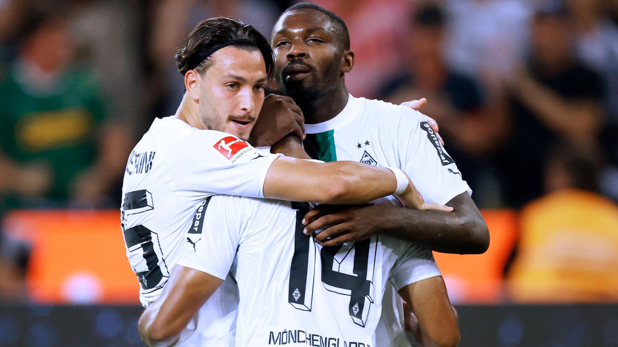Die Gladbach-Profis Ramy Bensebaini (l.) und Marcus Thuram (r.) umarmen ihren Teamkollegen Alassane Plea, hier zu sehen am 19. August 2022 im Borussia-Park. Von Plea ist die Rückseite zu sehen.