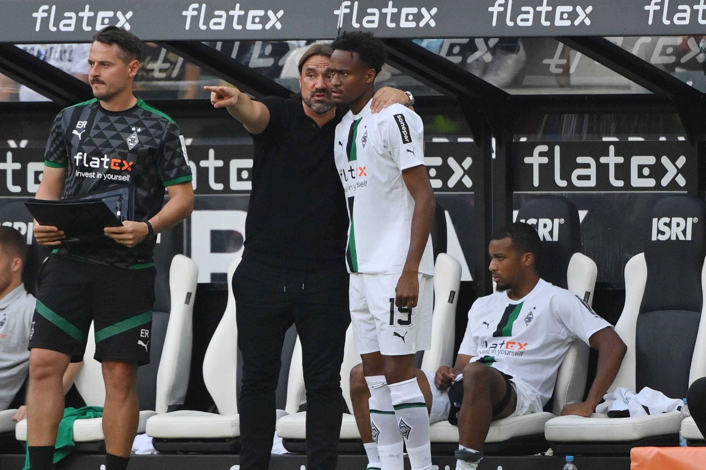 Trainer Daniel Farke von Borussia Mönchengladbach gibt Nathan Ngoumou Anweisung vor dessen Einwechslung am 4. September 2022 gegen Mainz 05. Farke hat den Arm um Ngoumous Schulter gelegt und zeigt mit dem anderen Arm auf das Spielfeld. Links steht Co-Trainer Edmund Riemer, im Hintergrund sitzt Alassane Plea auf der Bank.
