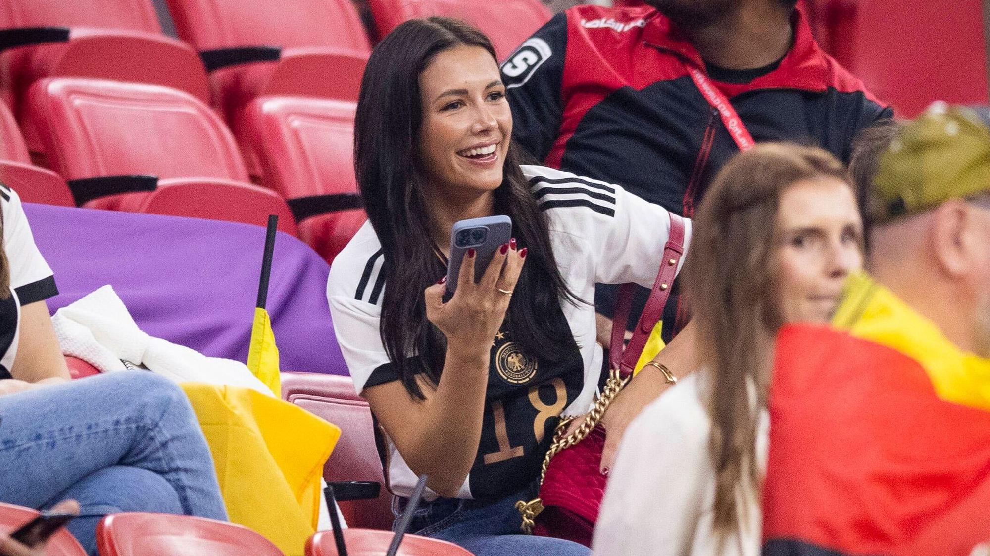 Sky-Moderatorin Laura Winter, hier zu sehen am 1. Dezember 2022 während des WM-Spiels der deutschen Nationalmannschaft gegen Costa Rica in Doha, ist mit DFB-Spieler und Gladbach-Star Jonas Hofmann verlobt. Winter trägt ein Deutschland-Trikot und hält ein Smartphone in ihrer rechte Hand. Sie lächelt zudem.