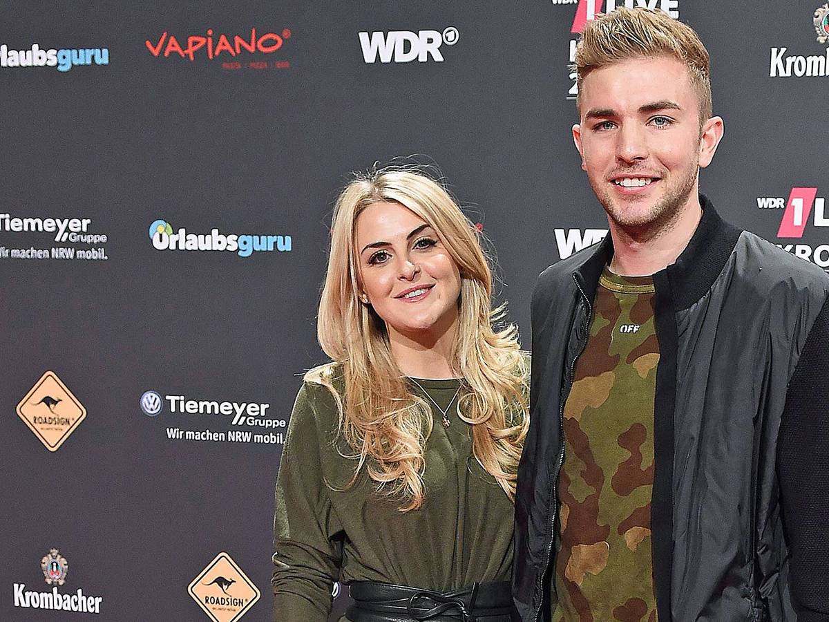 ZDF-Experte und Gladbach-Profi Christoph Kramer (r.) mit seiner Frau Celina (l.) bei der „1LIVE Krone“ am 7. Dezember 2017 in der Jahrhunderthalle in Bochum. Die beiden lächeln, hinter ihnen ist eine Werbewand zu sehen.