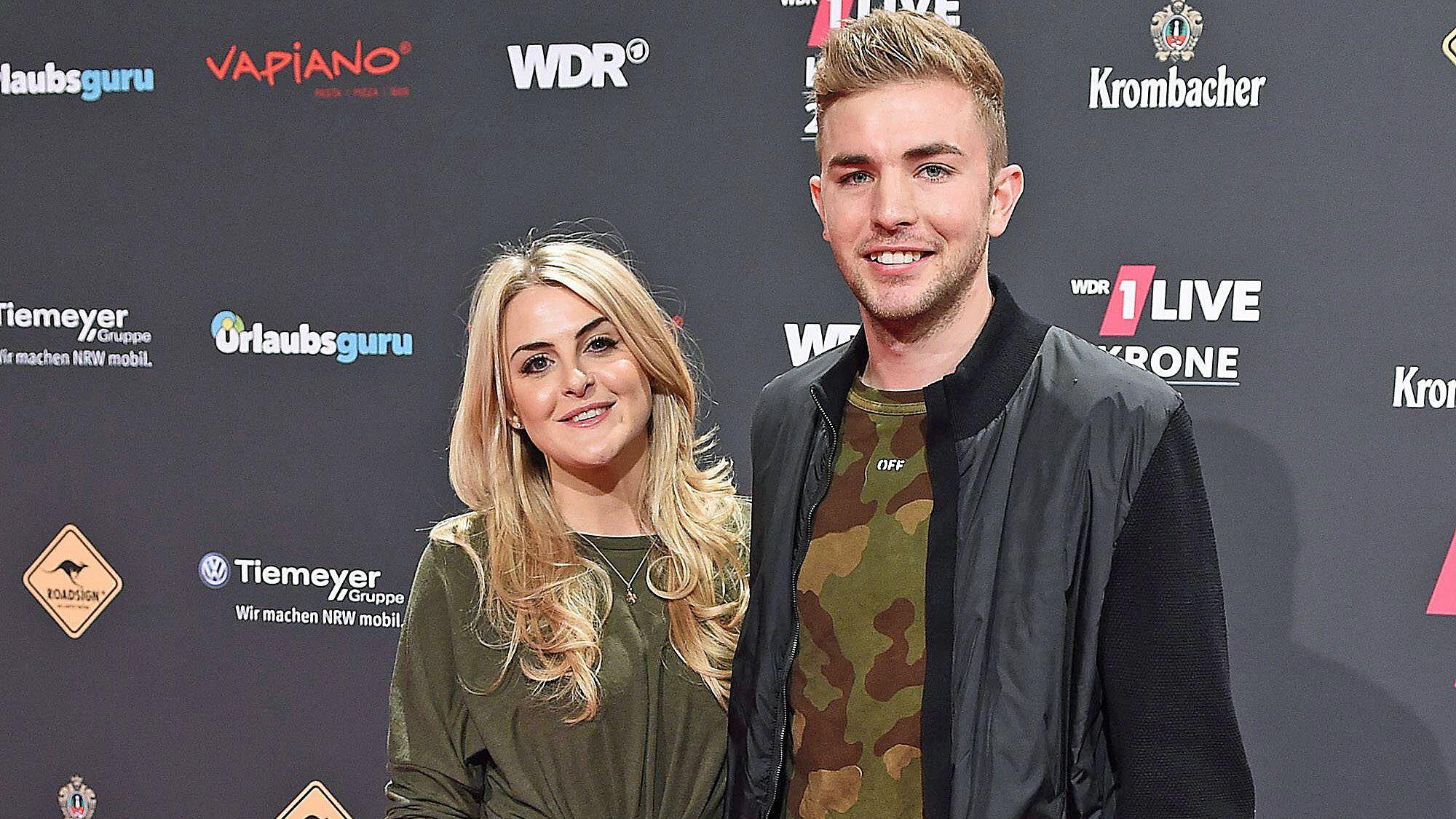 ZDF-Experte und Gladbach-Profi Christoph Kramer (r.) mit seiner Frau Celina (l.) bei der „1LIVE Krone“ am 7. Dezember 2017 in der Jahrhunderthalle in Bochum. Die beiden lächeln, hinter ihnen ist eine Werbewand zu sehen.