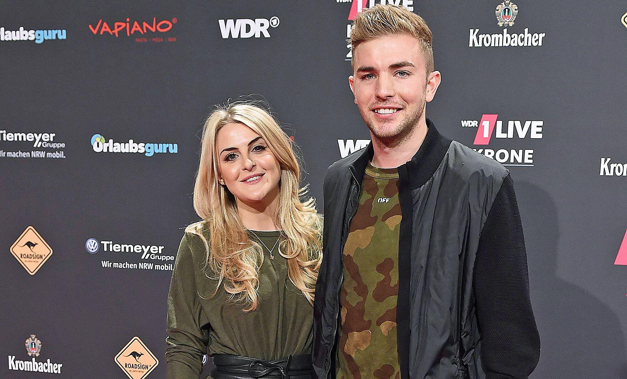 ZDF-Experte und Gladbach-Profi Christoph Kramer (r.) mit seiner Frau Celina (l.) bei der „1LIVE Krone“ am 7. Dezember 2017 in der Jahrhunderthalle in Bochum. Die beiden lächeln, hinter ihnen ist eine Werbewand zu sehen.