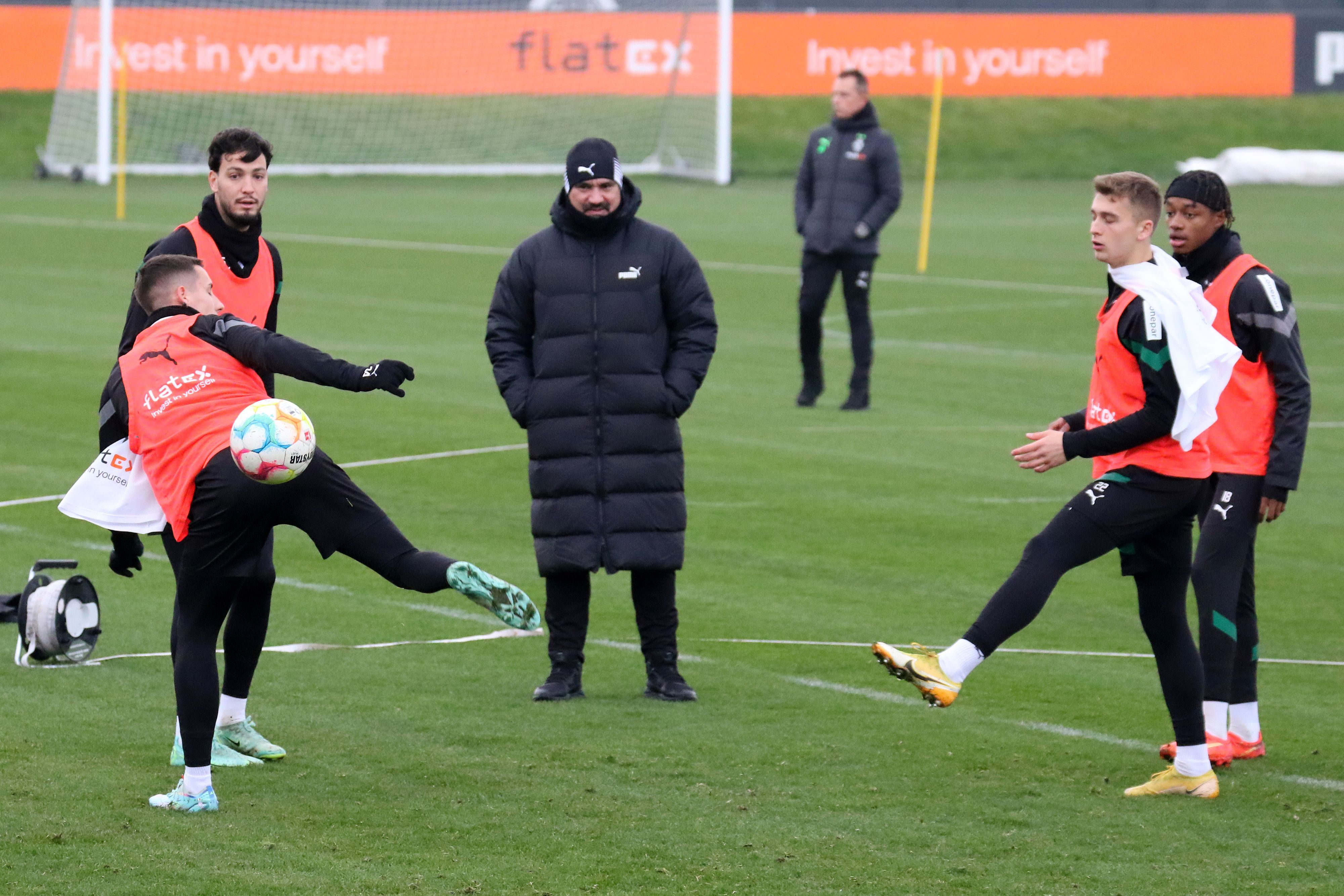 Trainings von Borussia Mönchengladbach im Borussia-Park am 22. Dezember 2022. Trainer Daniel Farke (M.) schaut seinen Spielern Conor Noß, Ramy Bensebaini, Oscar Fraulo und Yvandro Borges Sanches (v. l. n. r.) zu. Farke trägt eine lange Winterjacke und eine Mütze.