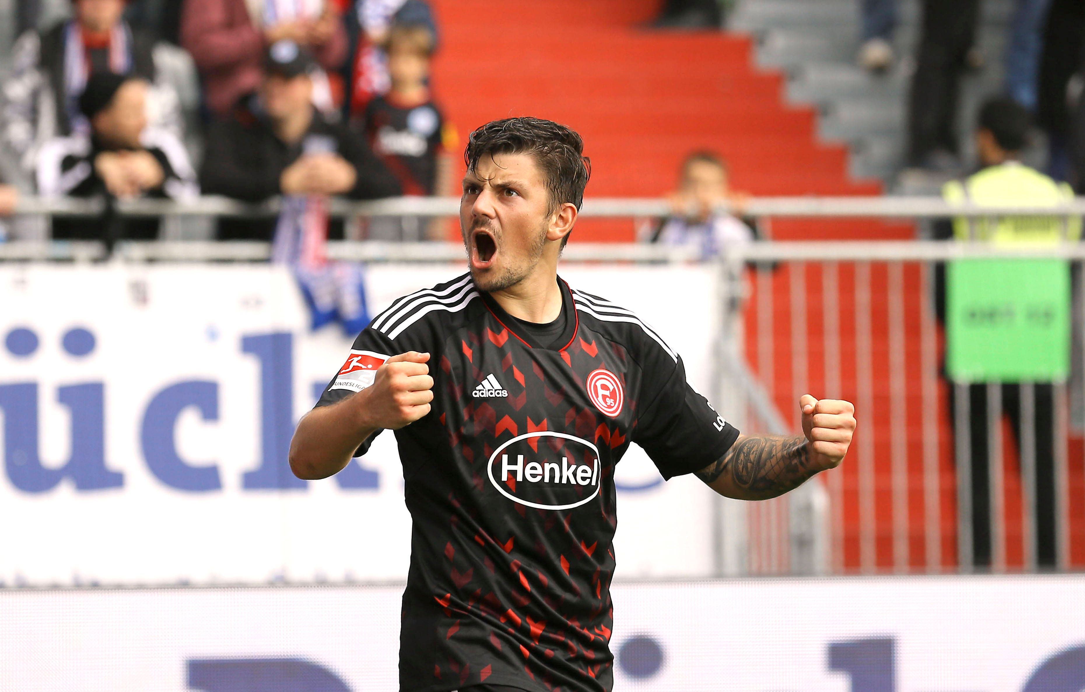 Dawid Kownacki von Fortuna Düsseldorf brüllt nach einem Tor den Fans auf der Tribüne zu und jubelt mit nach vorne gestreckten Armen. Der polnische Stürmer trägt das rot-schwarze Auswärtstrikot von Fortuna.