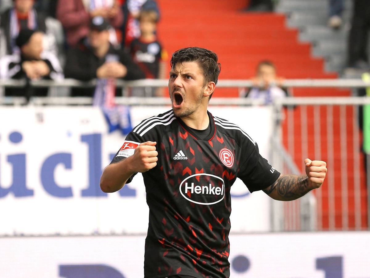 Dawid Kownacki von Fortuna Düsseldorf brüllt nach einem Tor den Fans auf der Tribüne zu und jubelt mit nach vorne gestreckten Armen. Der polnische Stürmer trägt das rot-schwarze Auswärtstrikot von Fortuna.