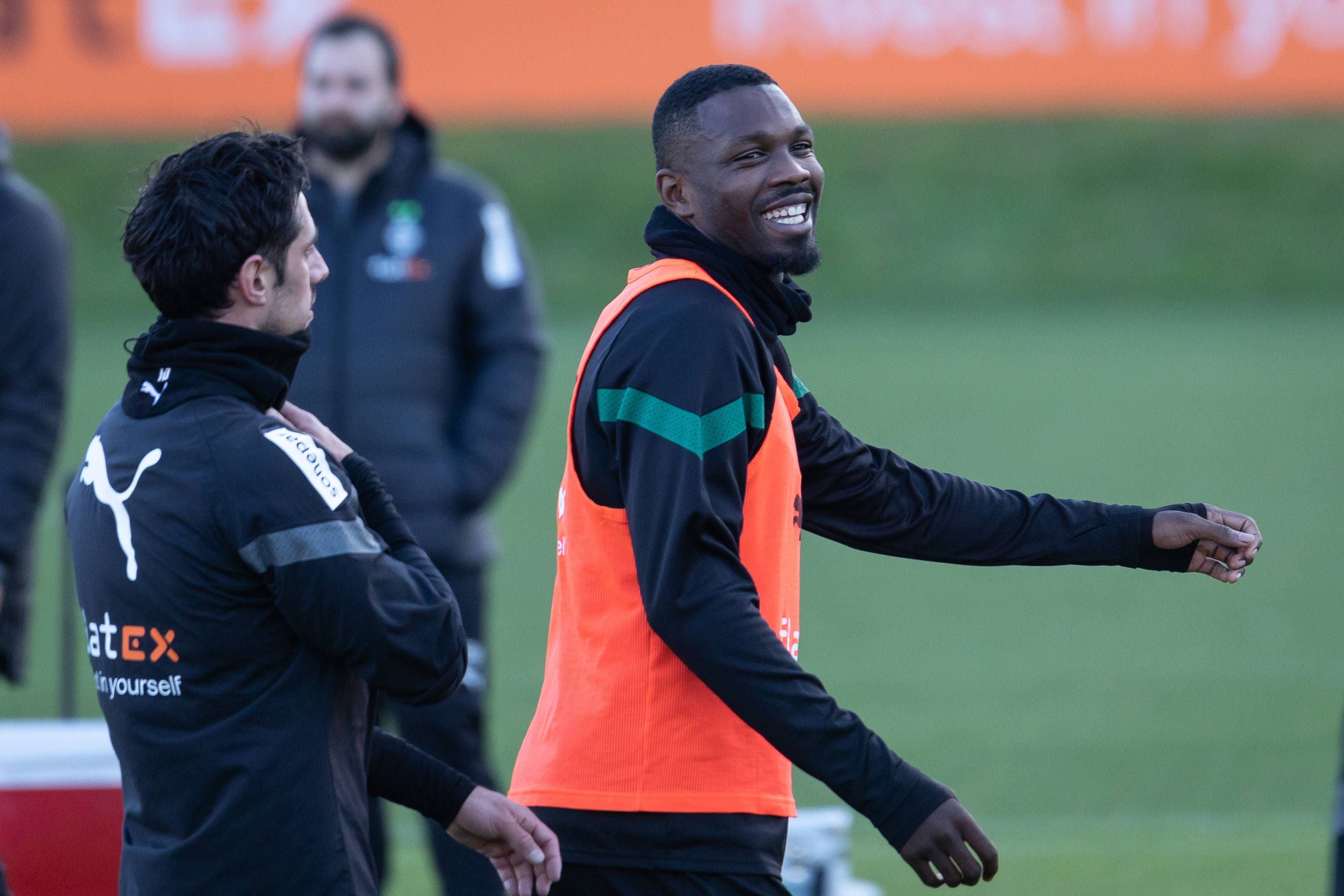 Marcus Thuram steht in Trainingskleidung und einem roten Leibchen am 6. Januar 2023 auf dem Trainingsplatz von Borussia Mönchengladbach. Der Fußballer lacht nach einem Gespräch mit Borussia-Teamkollege Lars Stindl.