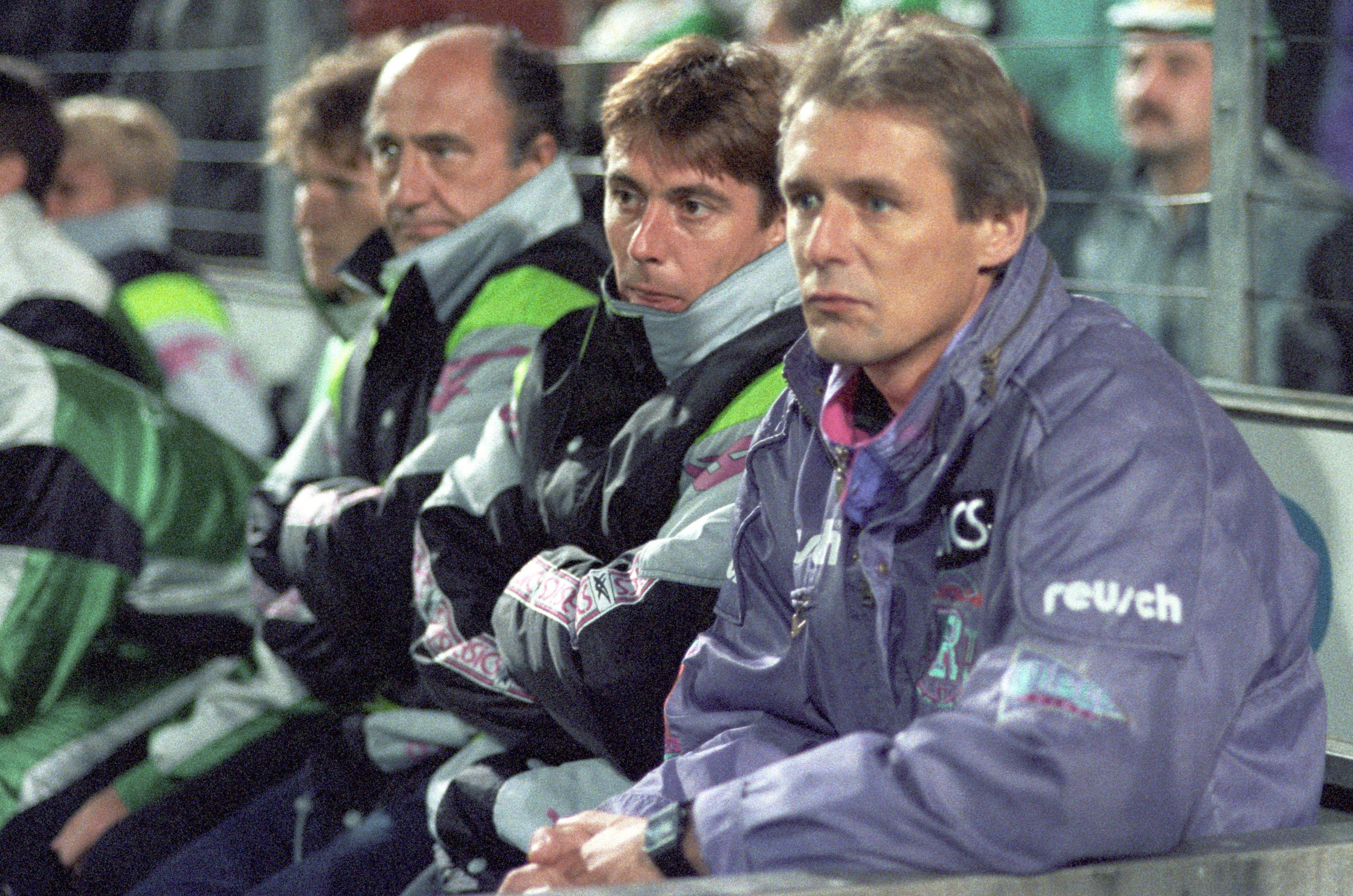 Jürgen Gelsdorf sitzt bei einem Spiel von Borussia Mönchengladbach im DFB-Pokal auf der Trainerbank und trägt einen Trainingsanzug. Neben ihm sitzt Bernd Krauss, sein damaliger Co-Trainer, in Trainingsjacke und mit verschränkten Armen.