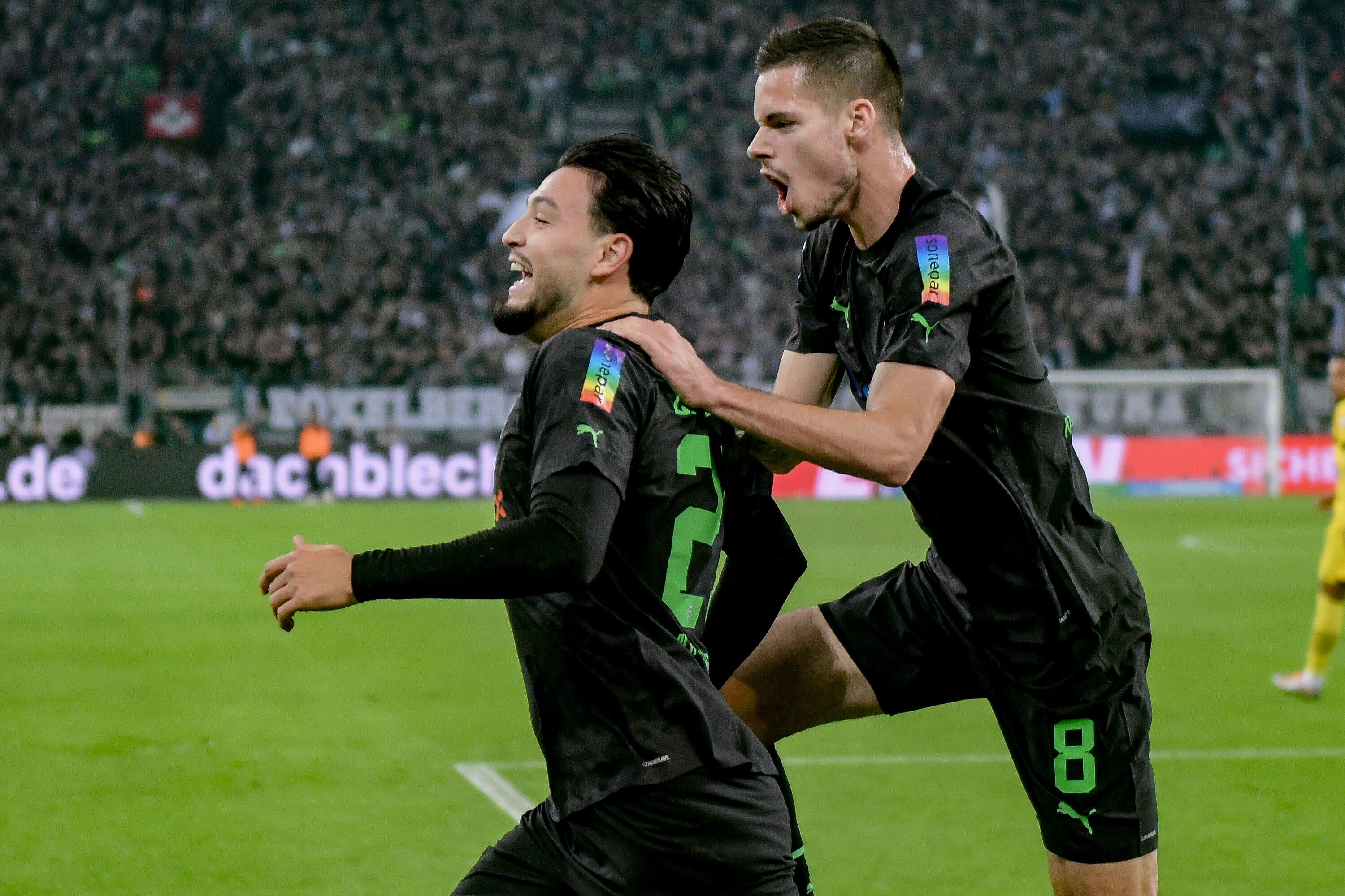 Julian Weigl (r.) hier am 11. November 2022 beim Torjubel mit Teamkollege Ramy Bensebaini (l.). Er springt ihm dabei auf den Rücken und schreit, Bensebaini grinst. Beide tragen das schwarze Gladbach-Trikot und sind von der Seite zu sehen.
