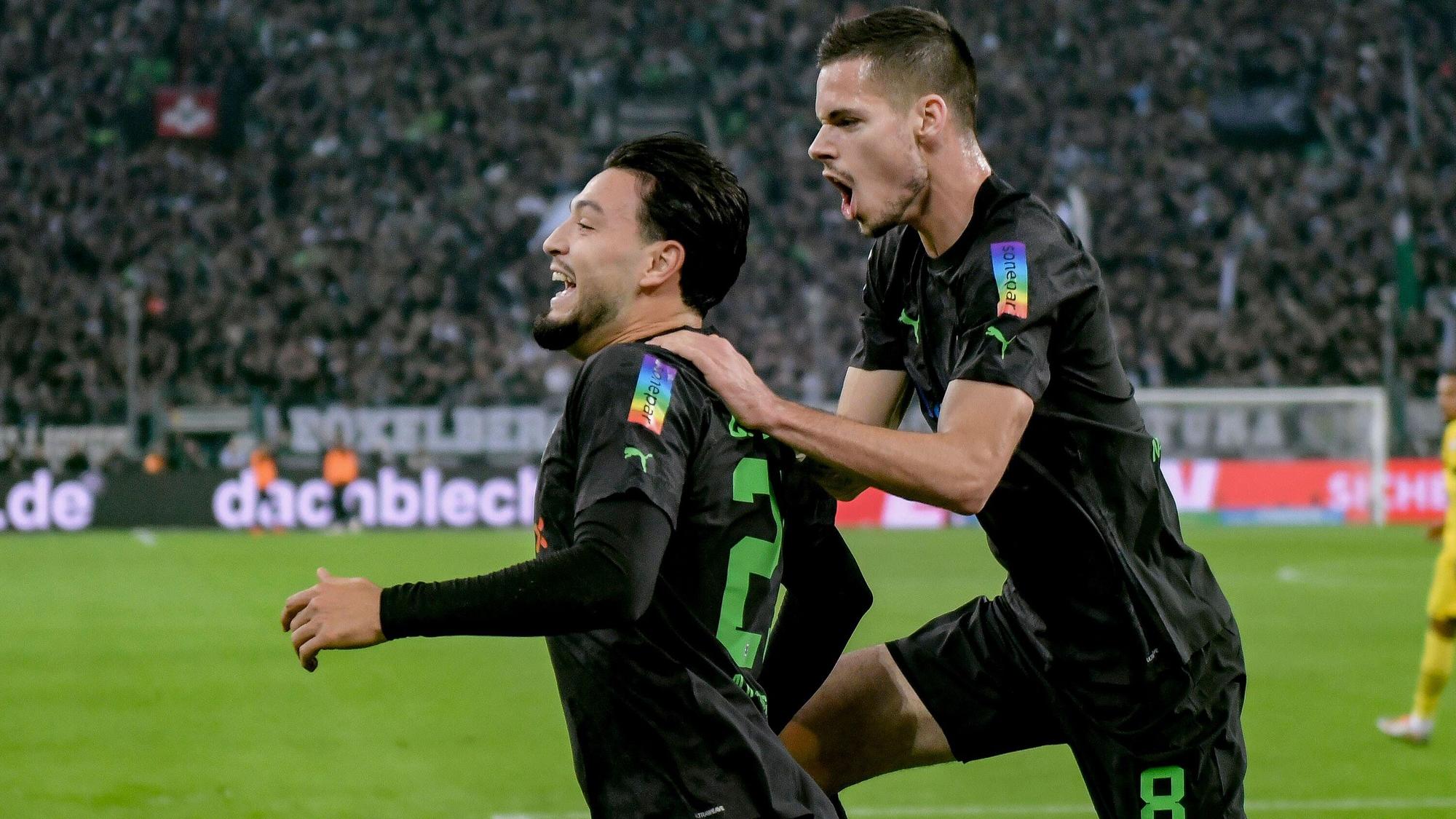 Julian Weigl (r.) hier am 11. November 2022 beim Torjubel mit Teamkollege Ramy Bensebaini (l.). Er springt ihm dabei auf den Rücken und schreit, Bensebaini grinst. Beide tragen das schwarze Gladbach-Trikot und sind von der Seite zu sehen.