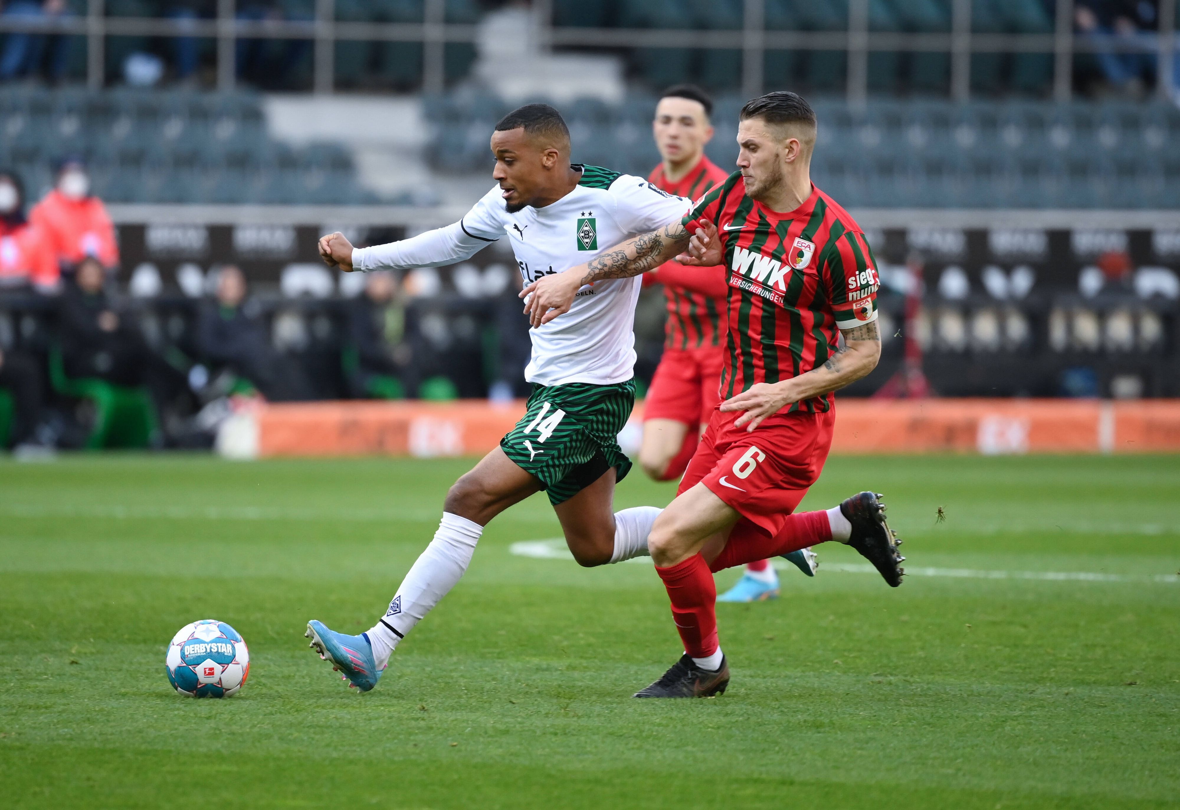 Alassane Plea (l.) von Borussia Mönchengladbach bestreitet einen Zweikampf mit Jeffrey Gouweleeuw vom FC Augsburg. Plea läuft dem Ball entgegen und trägt ein weiß-grünes Borussia-Trikot. Gouweleeuw verfolgt seinen Gegenspieler in einem rot-grünen Trikot.