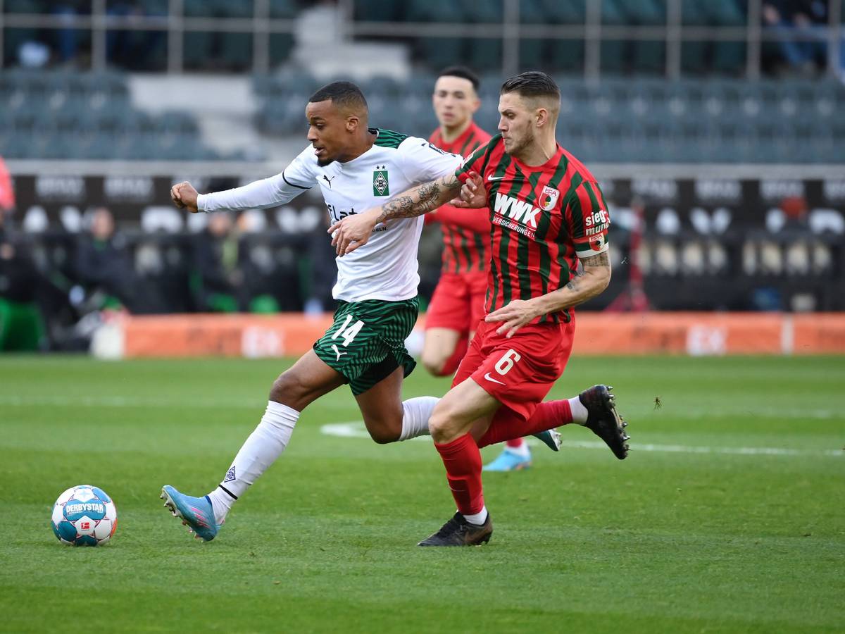 Alassane Plea (l.) von Borussia Mönchengladbach bestreitet einen Zweikampf mit Jeffrey Gouweleeuw vom FC Augsburg. Plea läuft dem Ball entgegen und trägt ein weiß-grünes Borussia-Trikot. Gouweleeuw verfolgt seinen Gegenspieler in einem rot-grünen Trikot.