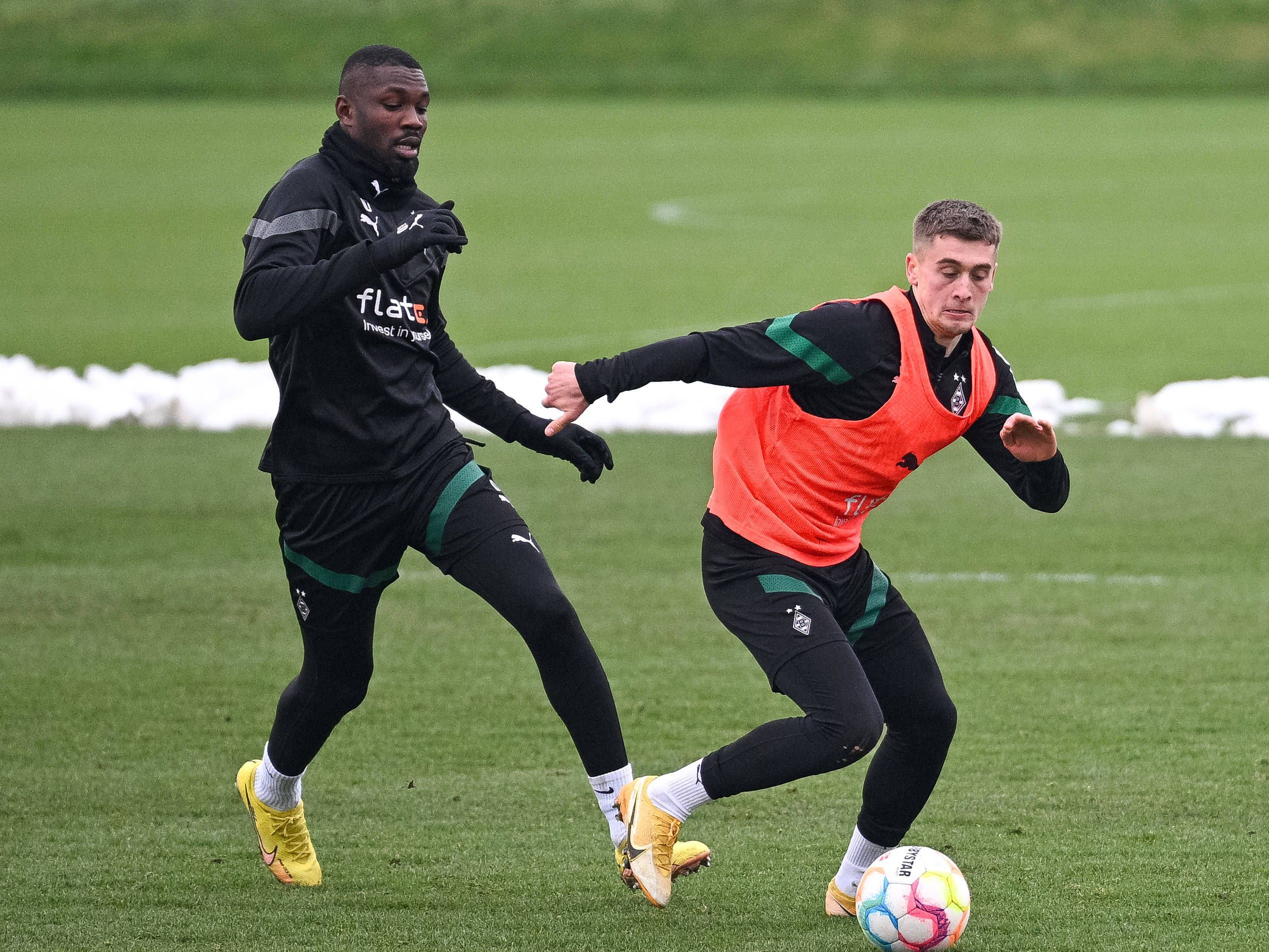 Gladbach-Stürmer Marcus Thuram (l.) im Duell mit Team-Kollege Oscar Fraulo (r.), hier zu sehen am Montag (23. Januar 2023) während einer Fohlen-Trainingseinheit im Borussia-Park. Fraulo trägt ein orangefarbenes Leibchen und hat den Ball vor sich.