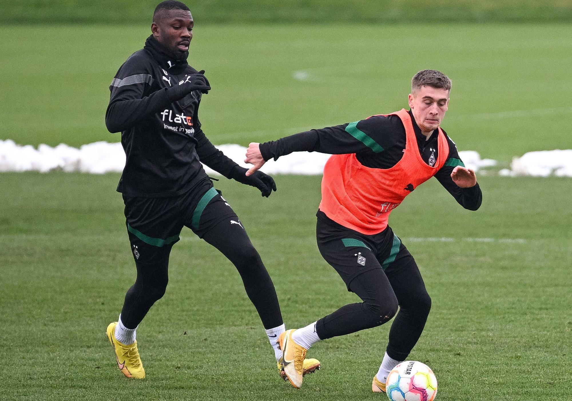 Gladbach-Stürmer Marcus Thuram (l.) im Duell mit Team-Kollege Oscar Fraulo (r.), hier zu sehen am Montag (23. Januar 2023) während einer Fohlen-Trainingseinheit im Borussia-Park. Fraulo trägt ein orangefarbenes Leibchen und hat den Ball vor sich.