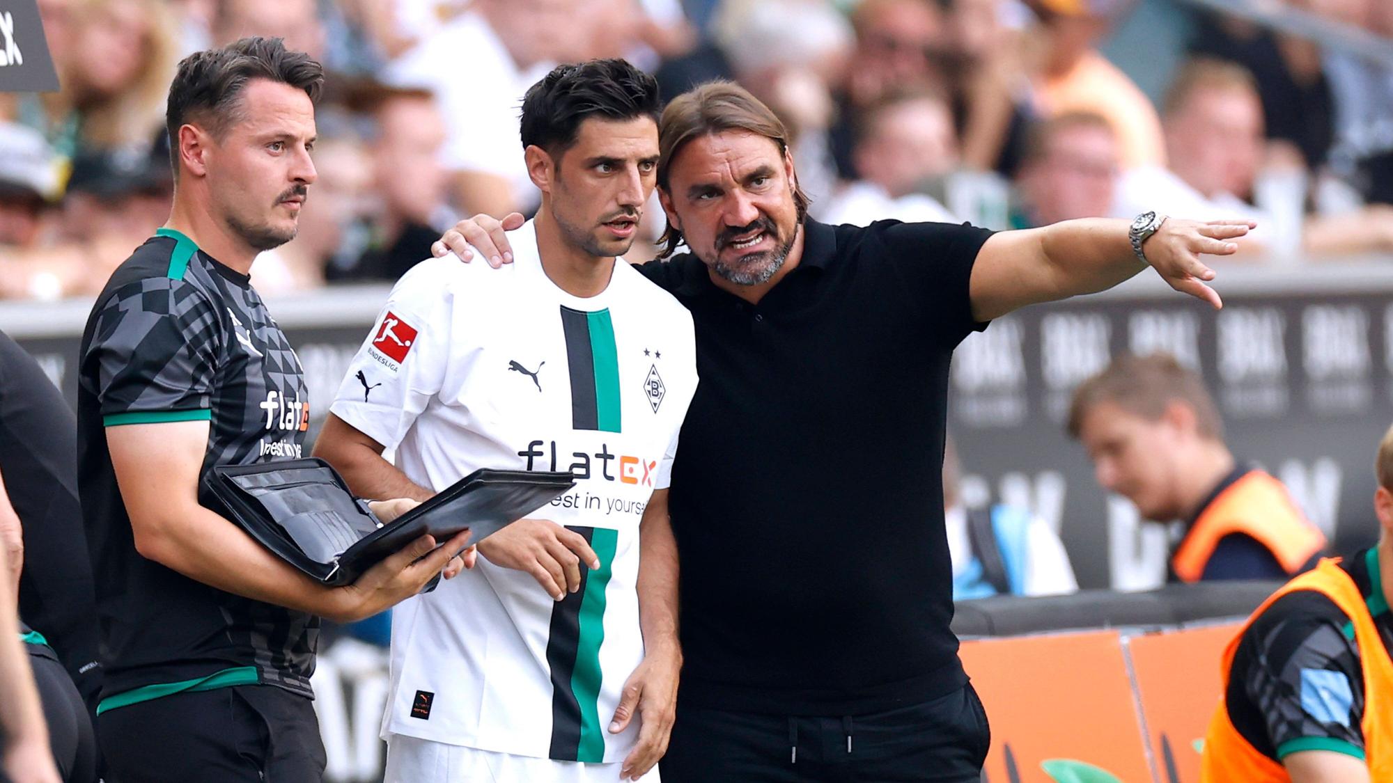 Daniel Farke (r.) legt seinen Arm um Lars Stindl (M.) und gibt dem Kapitän von Borussia Mönchengladbach vor einer Einwechslung taktische Anweisungen. Farke macht zudem eine Geste mit seinem linken Arm. Neben Farke und Stindl steht Co-Trainer Edmund Riemer und hält eine Mappe in der Hand.