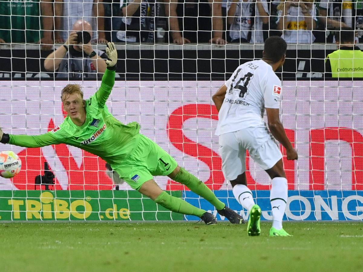 Alassane Plea steht nach einem geschossenen Elfmeter im Spiel von Borussia Mönchengladbach gegen Hertha BSC am 19. August 2022 vor Hertha-Torhüter Oliver Christensen, der dem Ball im rechten Eck hinterherspringt. Plea trägt ein weißes Gladbach-Trikot, Christensen das grüne Torhüter-Trikot von Hertha BSC.