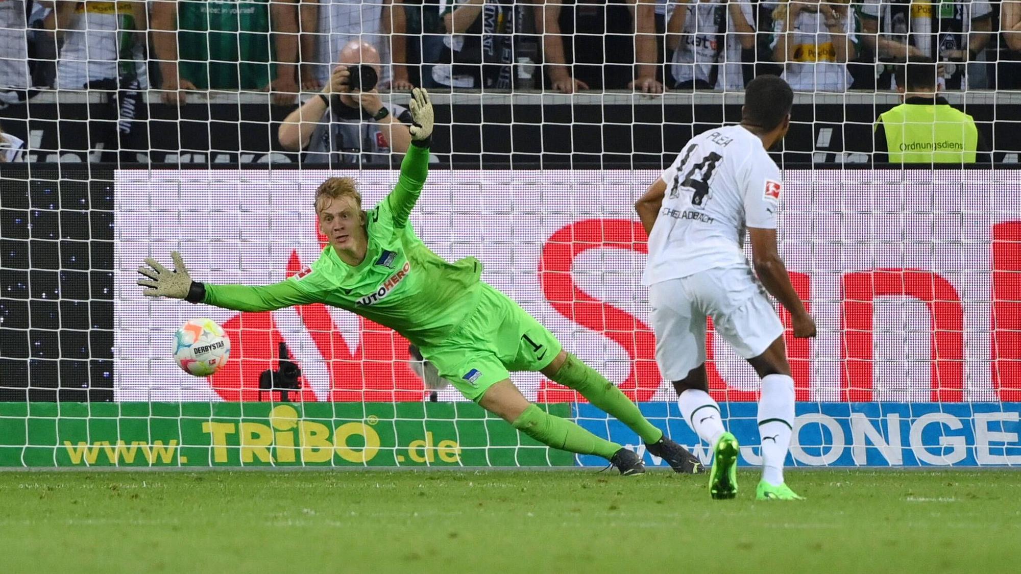 Alassane Plea steht nach einem geschossenen Elfmeter im Spiel von Borussia Mönchengladbach gegen Hertha BSC am 19. August 2022 vor Hertha-Torhüter Oliver Christensen, der dem Ball im rechten Eck hinterherspringt. Plea trägt ein weißes Gladbach-Trikot, Christensen das grüne Torhüter-Trikot von Hertha BSC.