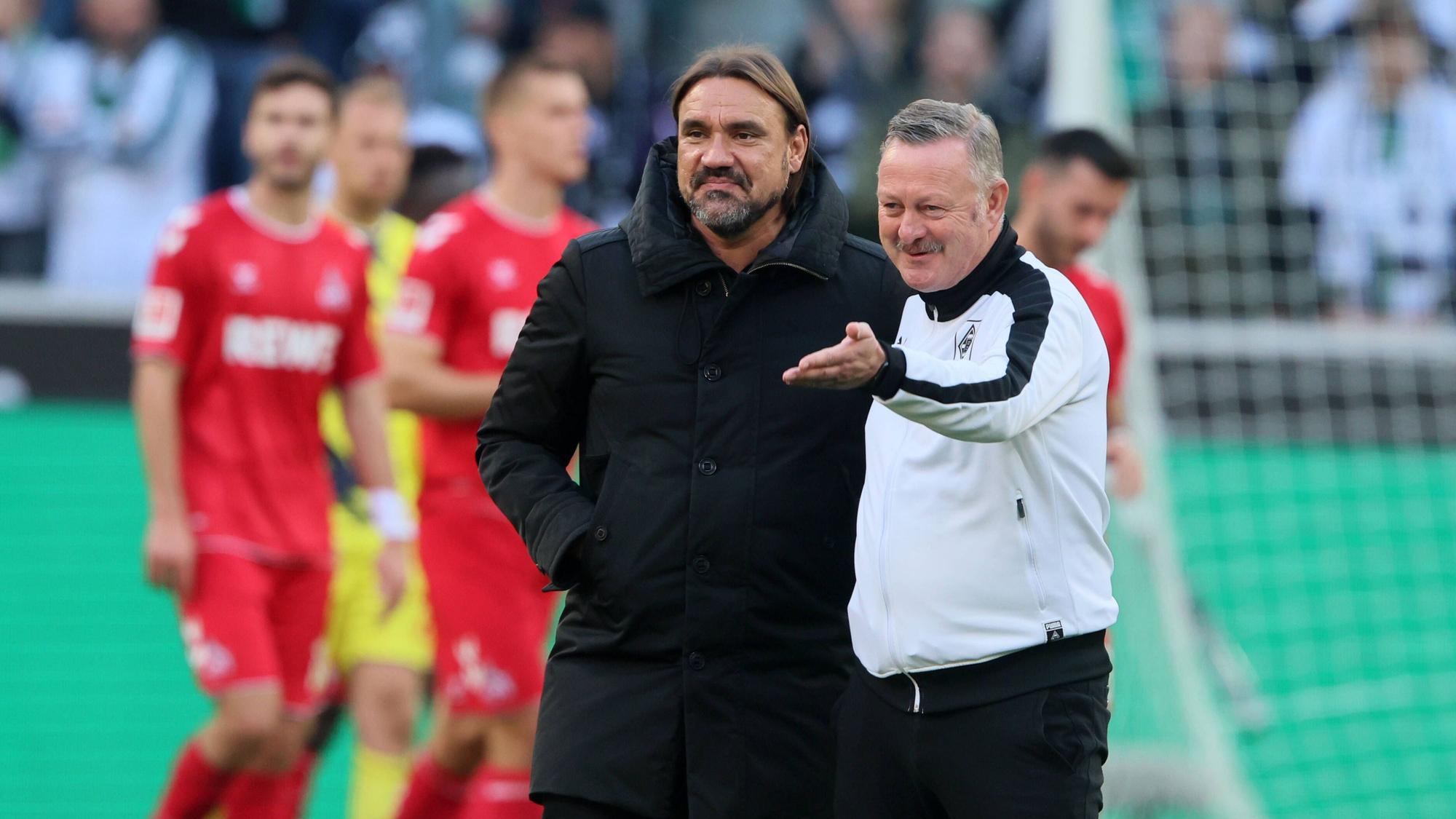 Daniel Farke (l.) und Roland Virkus stehen nach dem Bundesliga-Sieg im Derby gegen den 1. FC Köln auf dem Rasen im Borussia-Park nebeneinander und blicken in Richtung der Nordkurve. Virkus hebt seinen linken Arm und zeigt nach vorne.