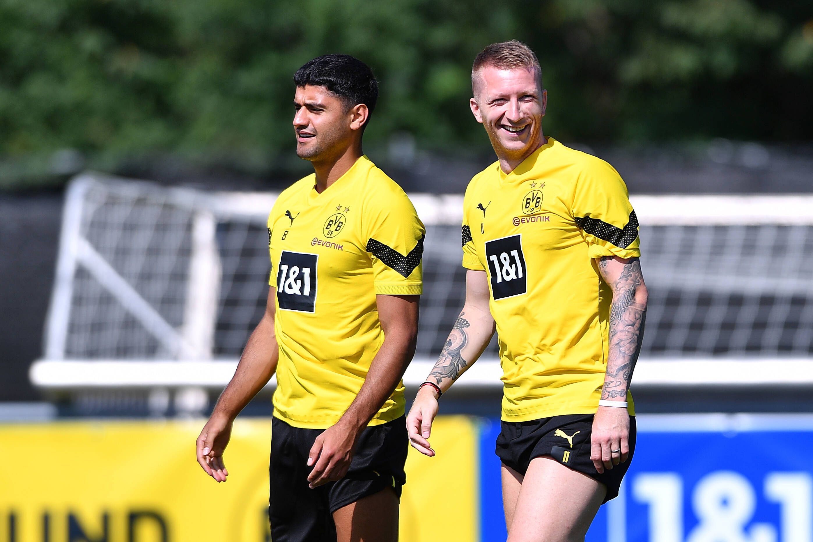 Mo Dahoud (l.) steht im Trainingslager von Borussia Dortmund neben seinem Teamkollegen Marco Reus. Beide tragen auf dem Rasen Trainingskleidung des BVB und lachen.