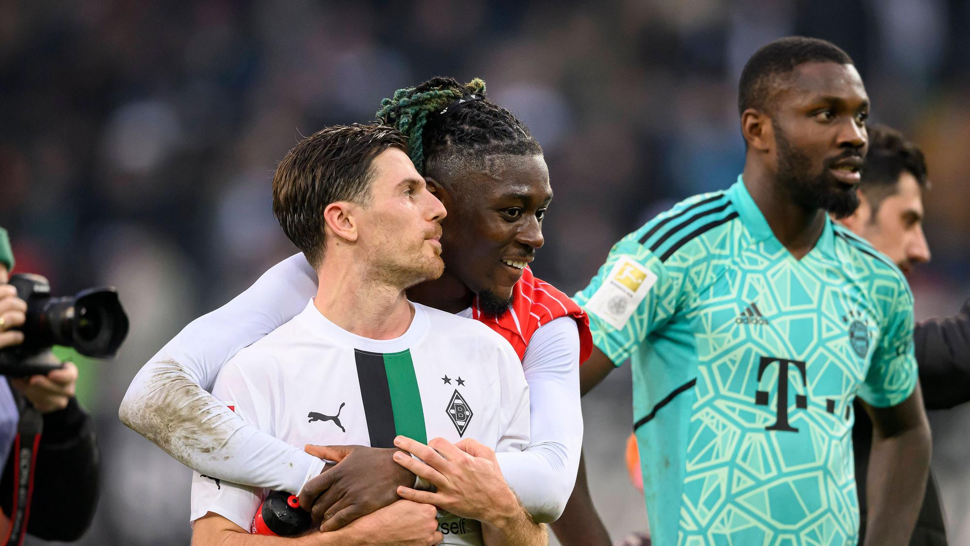 Manu Koné steht nach dem Bundesliga-Sieg von Borussia Mönchengladbach gegen den FC Bayern München am 18. Februar 2023 hinter seinem Teamkollegen Jonas Hofmann und legt seine Arme freundschaftlich um ihn. Marcus Thuram (r.) steht im Bayern-Trikot von Yann Sommer neben dem Gladbach-Duo auf dem Rasen im Borussia-Park.