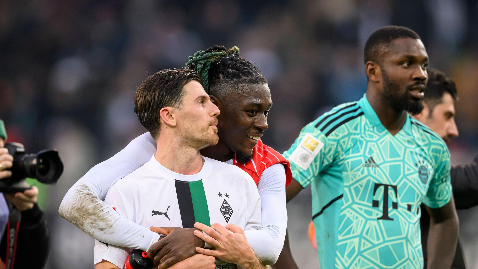 Manu Koné steht nach dem Bundesliga-Sieg von Borussia Mönchengladbach gegen den FC Bayern München am 18. Februar 2023 hinter seinem Teamkollegen Jonas Hofmann und legt seine Arme freundschaftlich um ihn. Marcus Thuram (r.) steht im Bayern-Trikot von Yann Sommer neben dem Gladbach-Duo auf dem Rasen im Borussia-Park.
