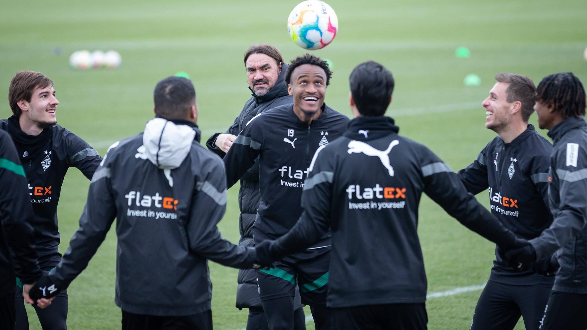 Ein bisschen Spaß muss sein! Gute Laune bei Borussia nach dem Heim-Coup gegen Bayern München. Die Gladbach-Profis, hier zu sehen am 16. Februar im Borussia-Park, beginnen nun mit der Vorbereitung auf das Mainz-Spiel. Zu sehen ist Ngoumou beim Kopfball, der Franzose lächelt, er hält seinen Team-Kollegen, die einen Kreis gebildet haben, an den Händen. Im Hintergrund schaut Trainer Daniel Farke zu.