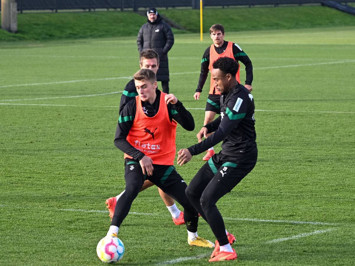 Kampf um den Ball zwischen den Gladbach-Talenten Oscar Fraulo (l.) und Nathan Ngoumou (r.) am 22. Dezember 2022 auf dem Trainingsgelände im Borussia-Park. Fraulo trägt ein Leibchen, beide Spieler sind auf den Ball fokussiert. Im Hintergrund schauen, Herrmann, Hofmann und Farke zu.