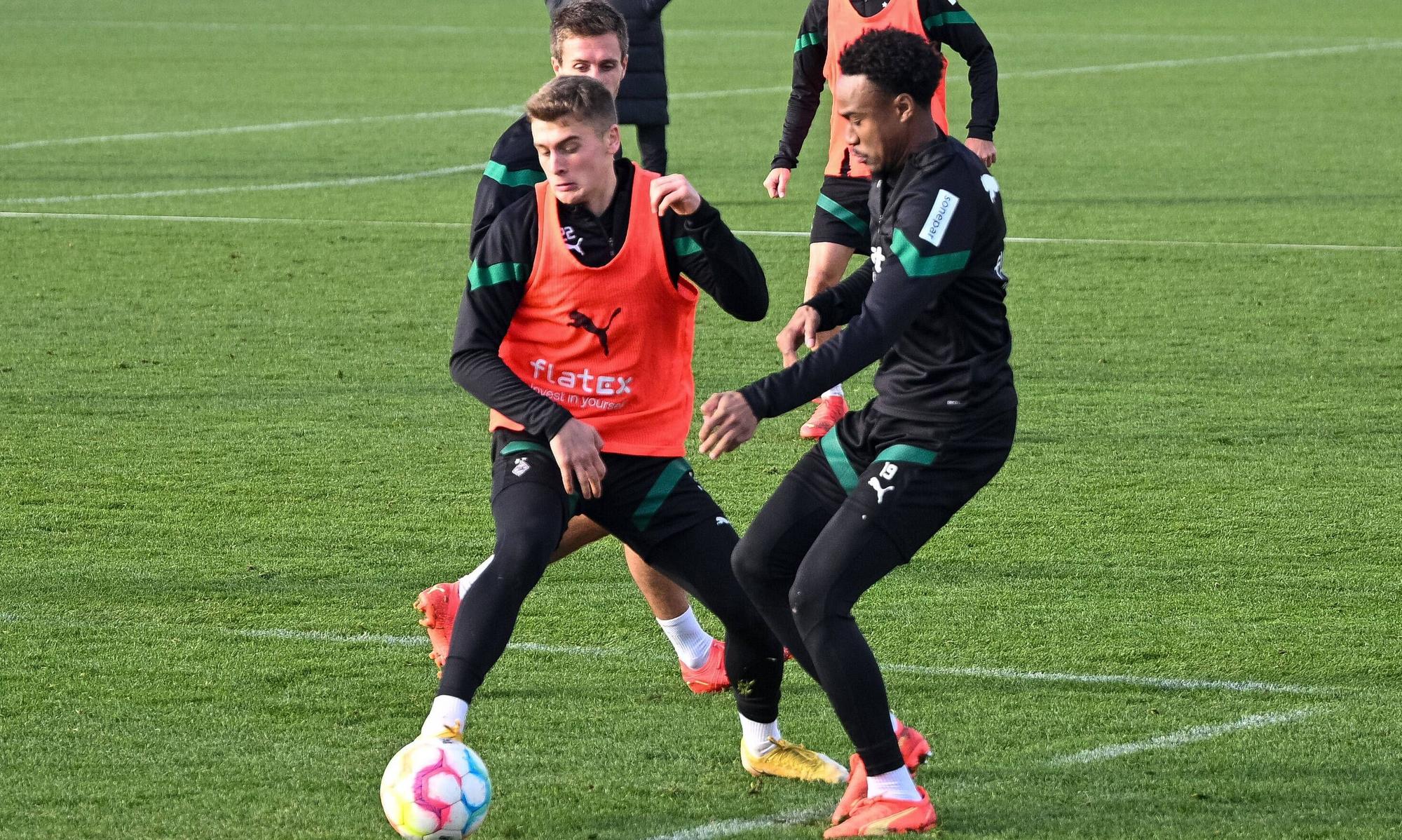Kampf um den Ball zwischen den Gladbach-Talenten Oscar Fraulo (l.) und Nathan Ngoumou (r.) am 22. Dezember 2022 auf dem Trainingsgelände im Borussia-Park. Fraulo trägt ein Leibchen, beide Spieler sind auf den Ball fokussiert. Im Hintergrund schauen, Herrmann, Hofmann und Farke zu.