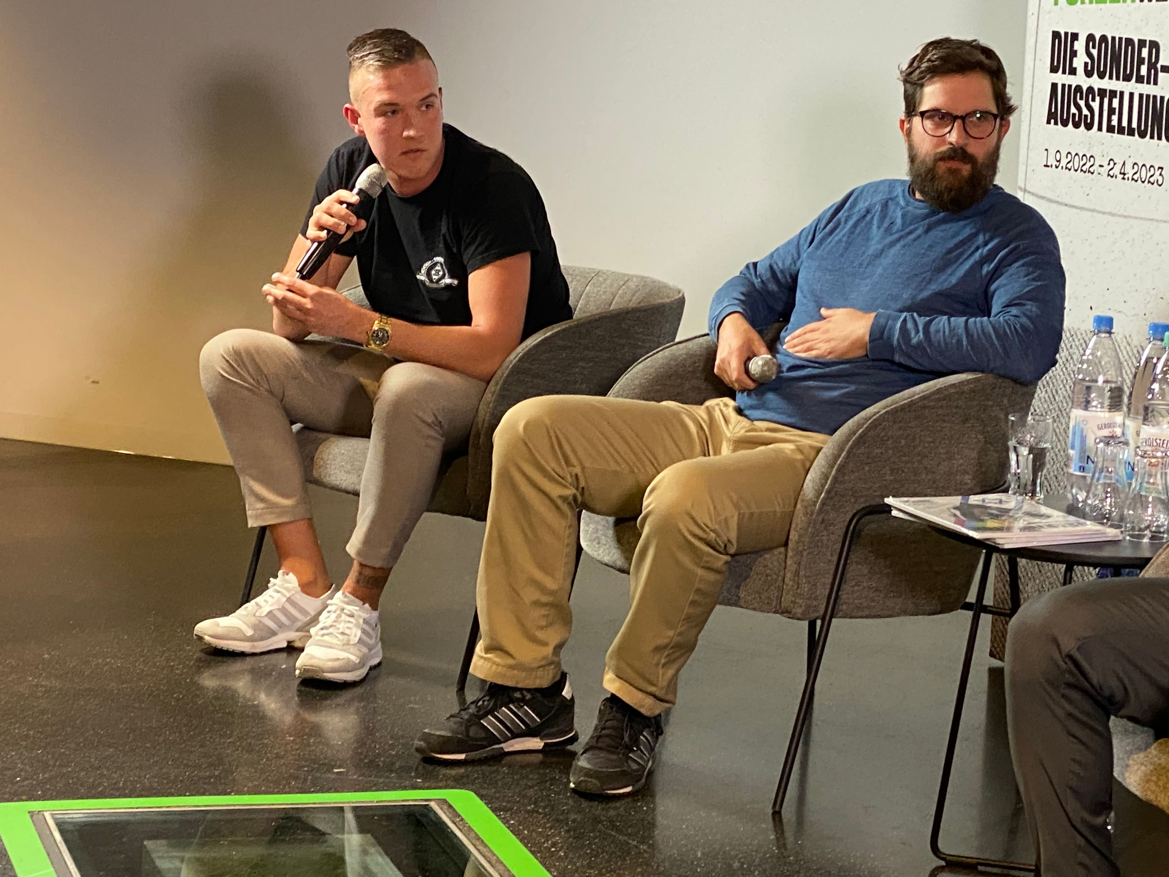 Simon Bender (l.) von der Gladbacher Ultra-Gruppierung „Sottocultura“ und der Politikwissenschaftler Jonas Gabler (r.) haben an der Diskussion (28. Februar 2023) mit Lars Stindl und Torsten Knippertz im Borussia-Park ebenfalls teilgenommen. Beide schauen zu Stindl rüber, Bender spricht in ein Mikrofon.