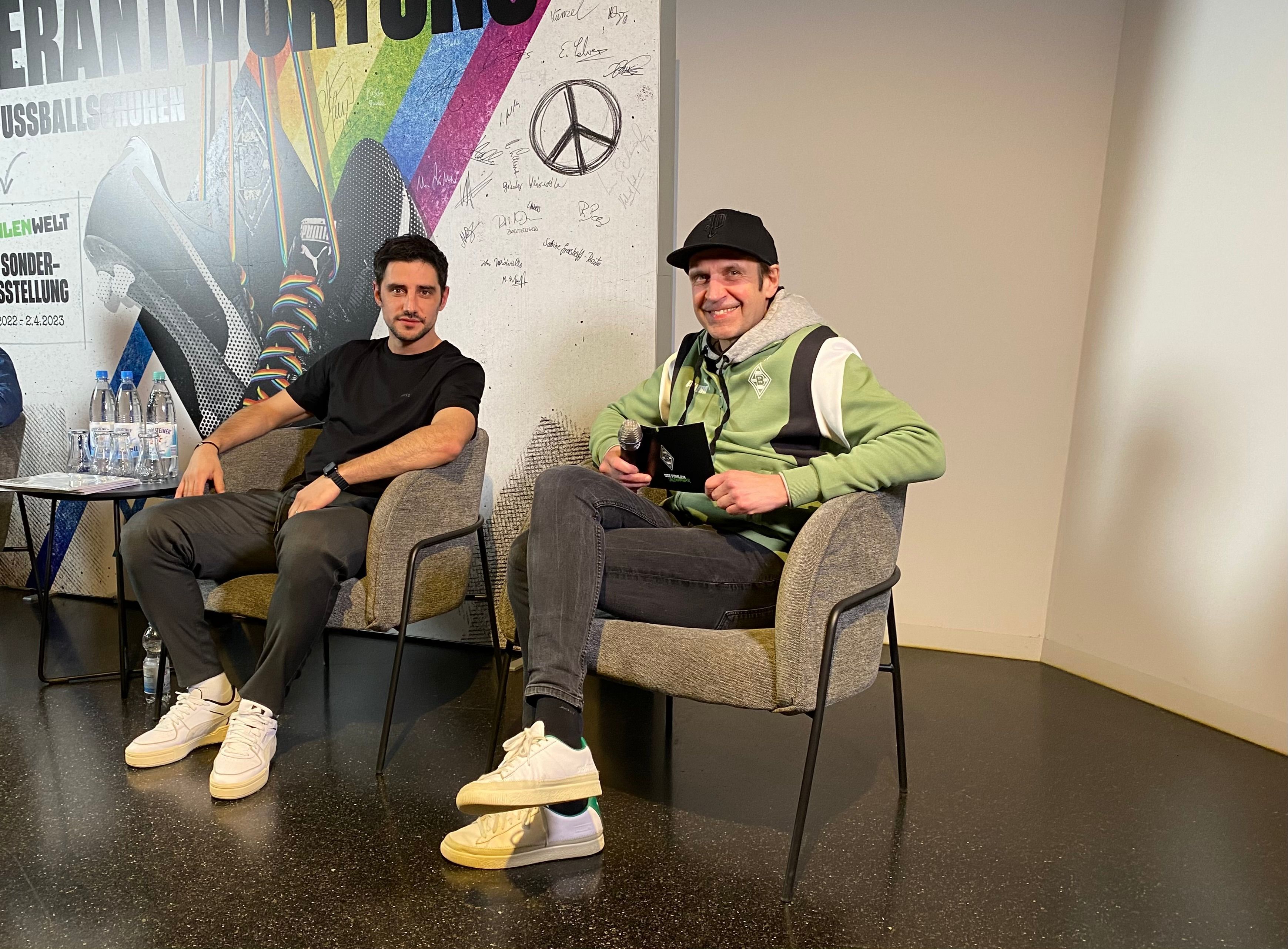 Gladbach-Kapitän Lars Stindl (l.) und Borussias Stadionsprecher Torsten Knippertz (r.) bei einer Diskussion (28. Februar 2023) zu den Themen Diskriminierung und Antidiskriminierung in der Fohlen-Welt im Borussia-Park. Beide schauen in die Kamera, Knippertz trägt eine Kappe.