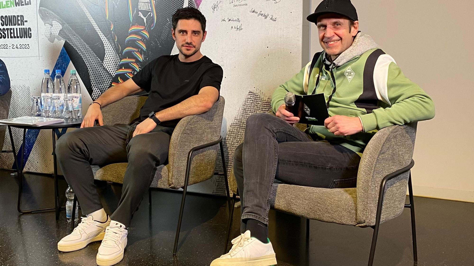 Gladbach-Kapitän Lars Stindl (l.) und Borussias Stadionsprecher Torsten Knippertz (r.) bei einer Diskussion (28. Februar 2023) zu den Themen Diskriminierung und Antidiskriminierung in der Fohlen-Welt im Borussia-Park. Beide schauen in die Kamera, Knippertz trägt eine Kappe.