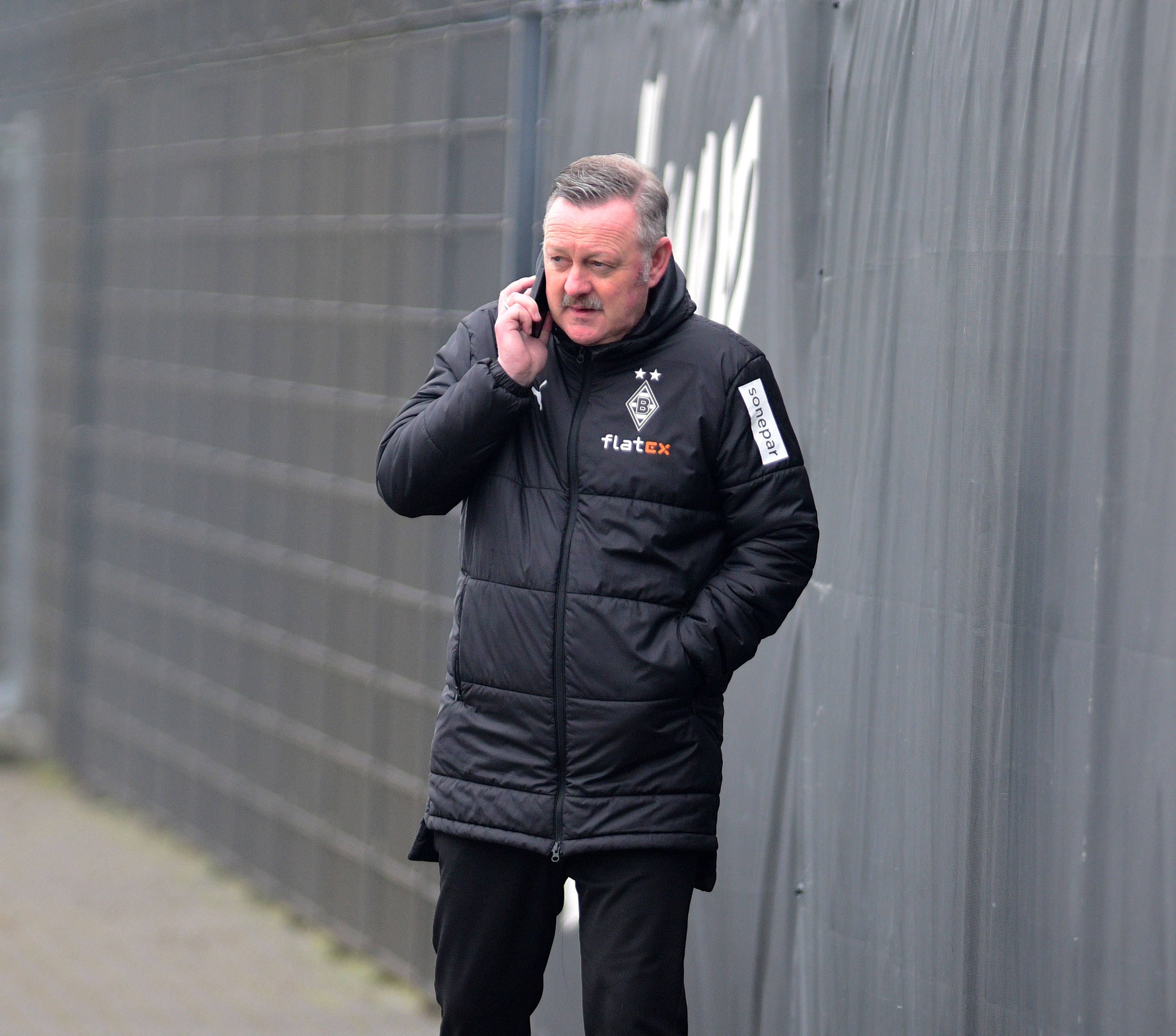 In Gesprächen: Borussia-Manager Roland Virkus telefoniert auf dem Trainingsplatz von Borussia Mönchengladbach während einer Einheit am 9. März 2023. Das Foto zeigt Virkus in einer Gladbach-Winterjacke und mit einem Telefon am rechten Ohr.