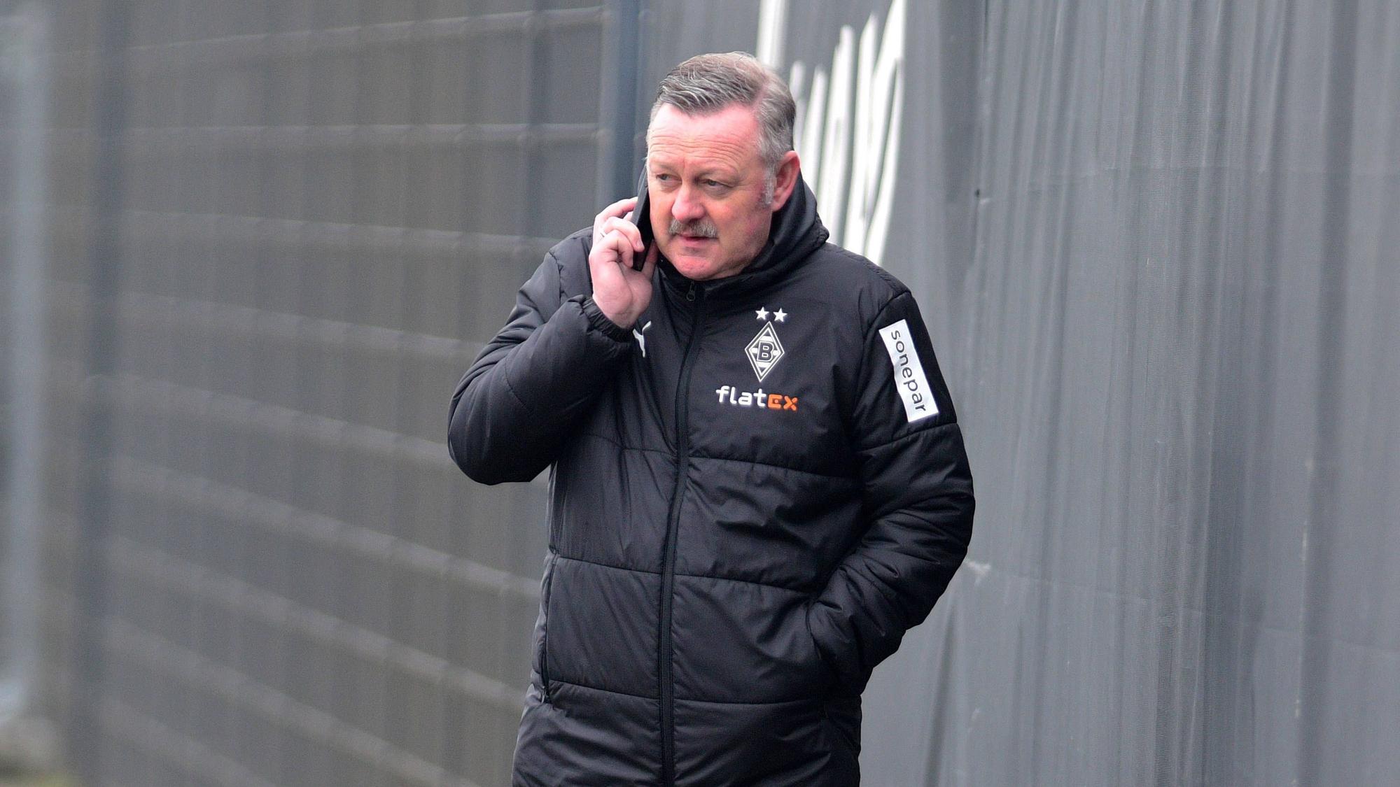 In Gesprächen: Borussia-Manager Roland Virkus telefoniert auf dem Trainingsplatz von Borussia Mönchengladbach während einer Einheit am 9. März 2023. Das Foto zeigt Virkus in einer Gladbach-Winterjacke und mit einem Telefon am rechten Ohr.