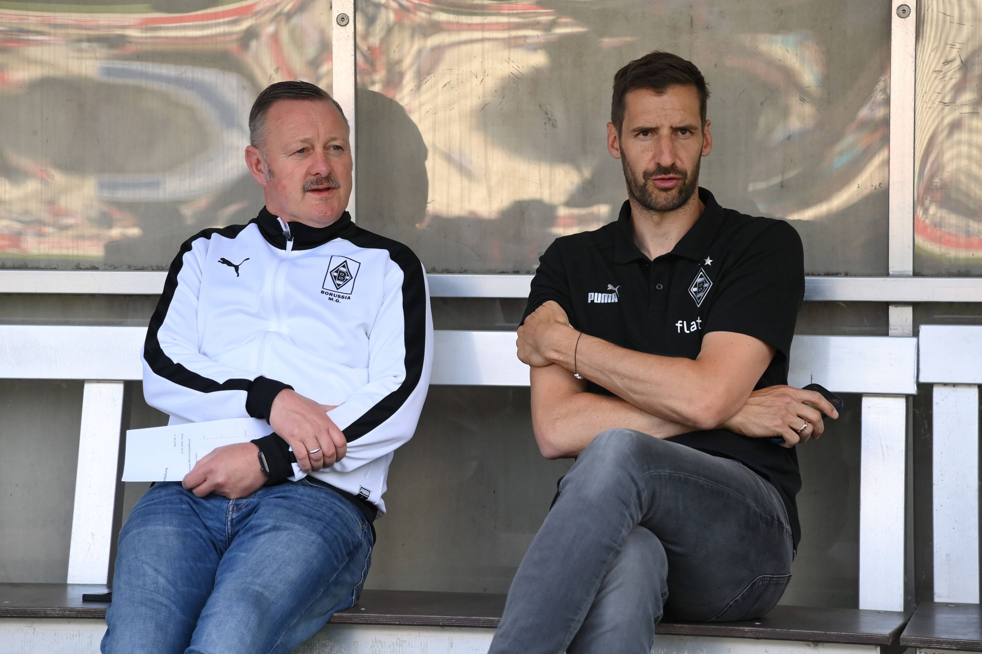Team-Manager Christofer Heimeroth (r.), hier am 1. Juli 2022 mit Fohlen-Manager Roland Virkus (l.) zu sehen, steht vor seinem Abschied bei Fußball-Bundesligist Borussia Moenchengladbach. Die beiden sitzen auf der Ersatzbank und schauen Richtung Spielfeld.