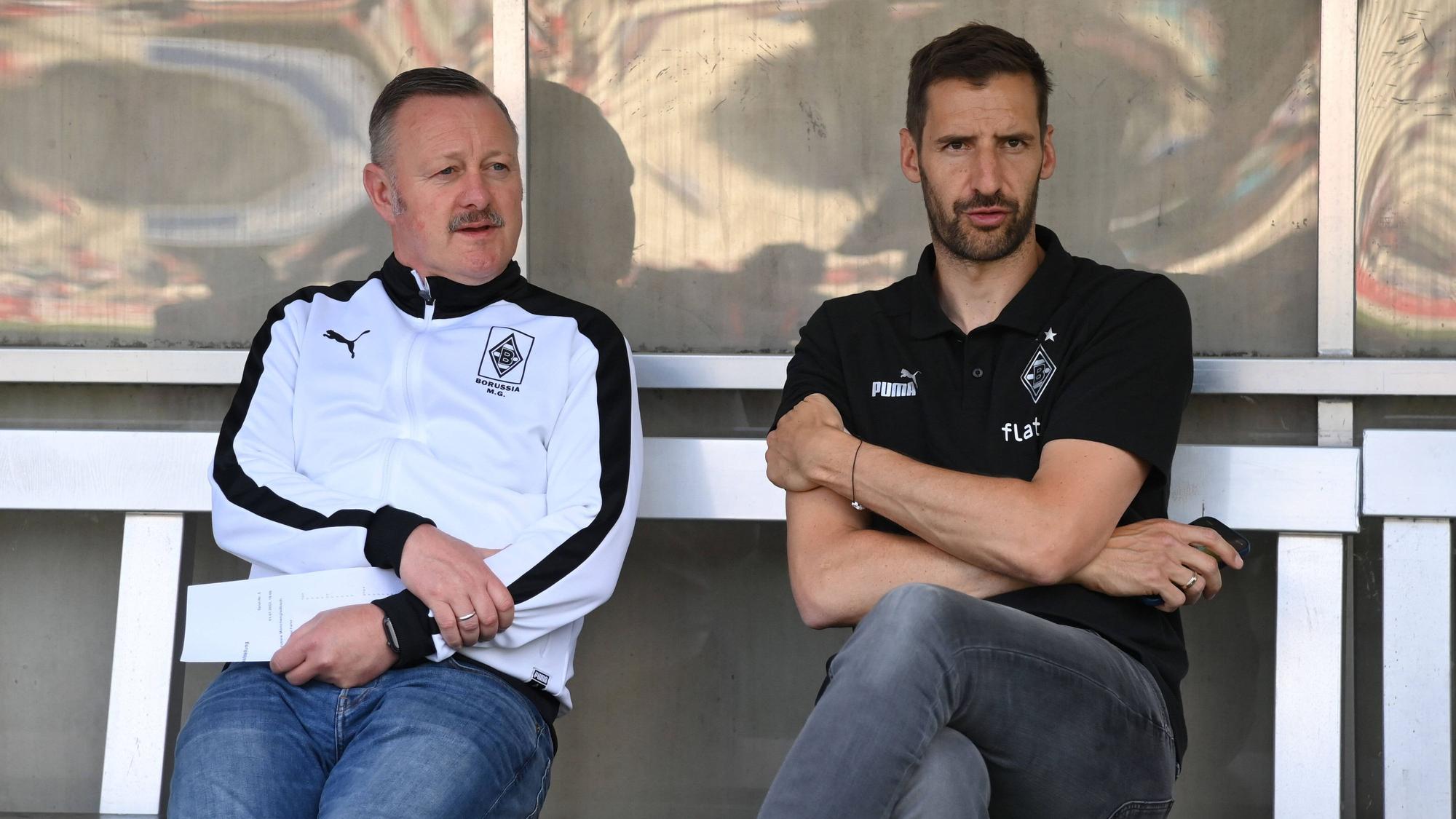 Team-Manager Christofer Heimeroth (r.), hier am 1. Juli 2022 mit Fohlen-Manager Roland Virkus (l.) zu sehen, steht vor seinem Abschied bei Fußball-Bundesligist Borussia Moenchengladbach. Die beiden sitzen auf der Ersatzbank und schauen Richtung Spielfeld.