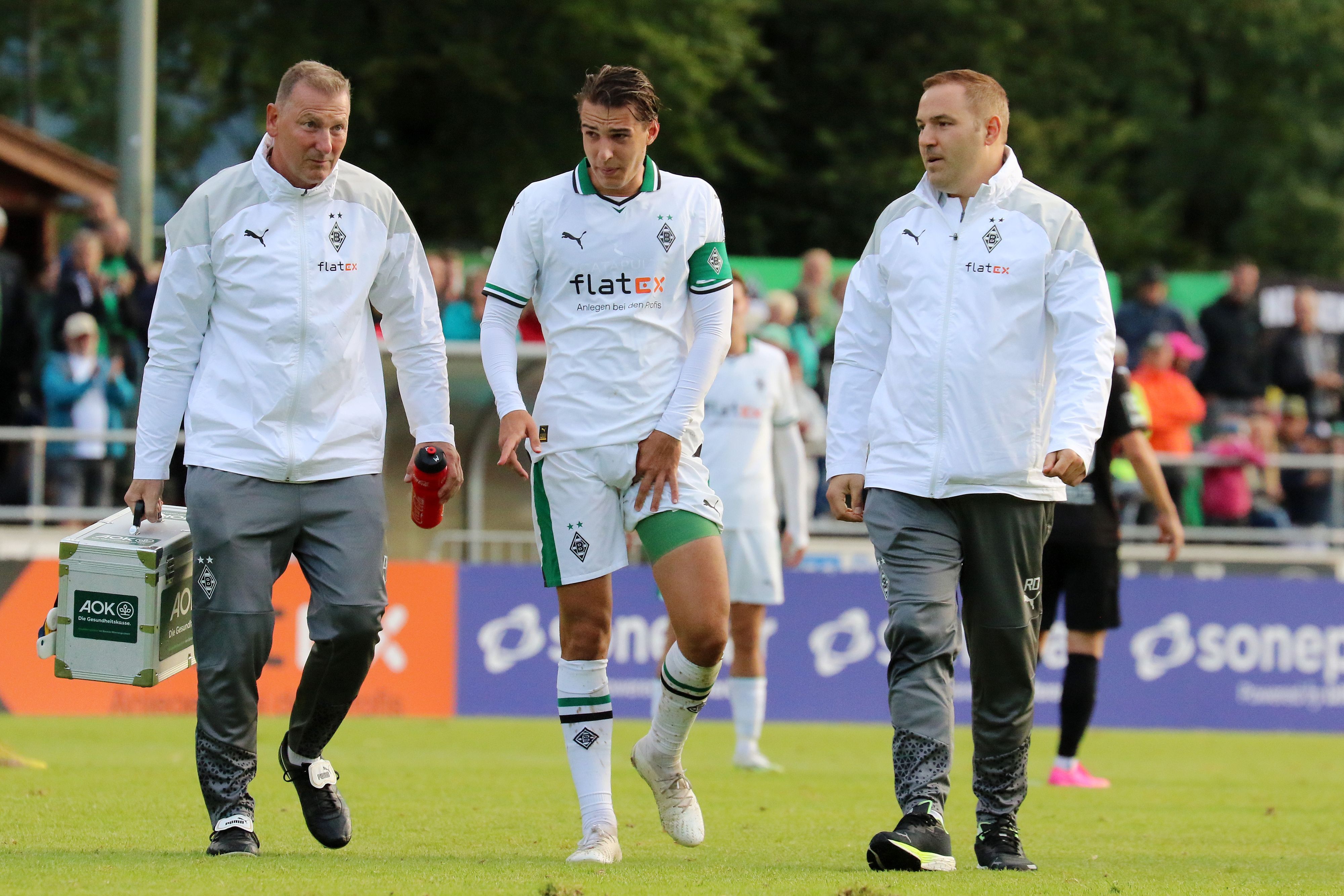 Florian Neuhaus (M.) verlässt gemeinsam mit Physiotherapeut Dirk Müller (l.) und Mannschaftsarzt Dr. Ralf Doyscher beim Testspiel von Borussia Mönchengladbach gegen den FC Ingolstadt am 26. Juli 2023 den Platz. Neuhaus' linker Oberschenkel ist bandagiert und er hält sich die Adduktoren.